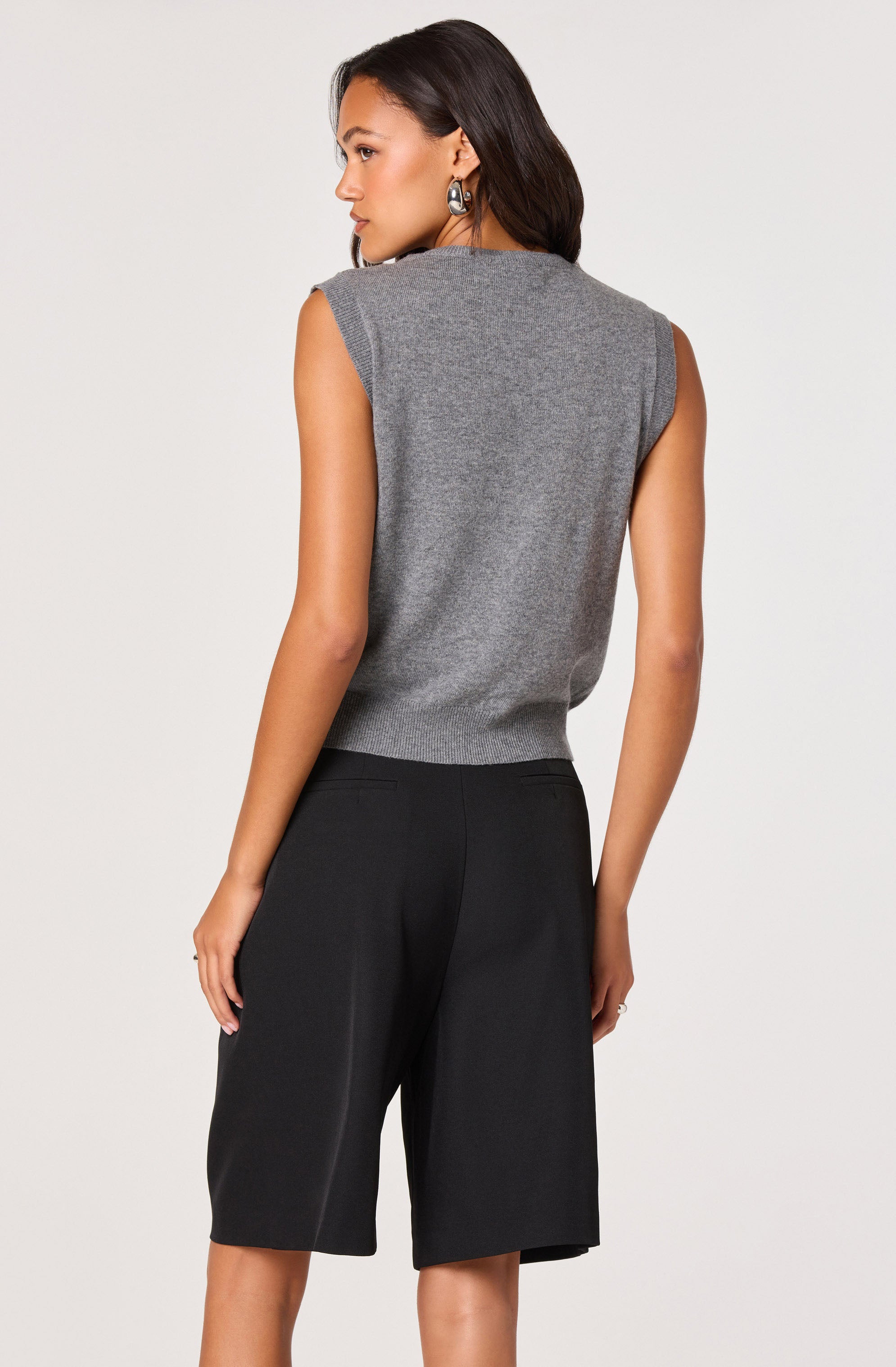 A woman is wearing a sleeveless, grey knit vest with a crew neckline and ribbed hem, paired with loose-fitting black knee-length shorts.