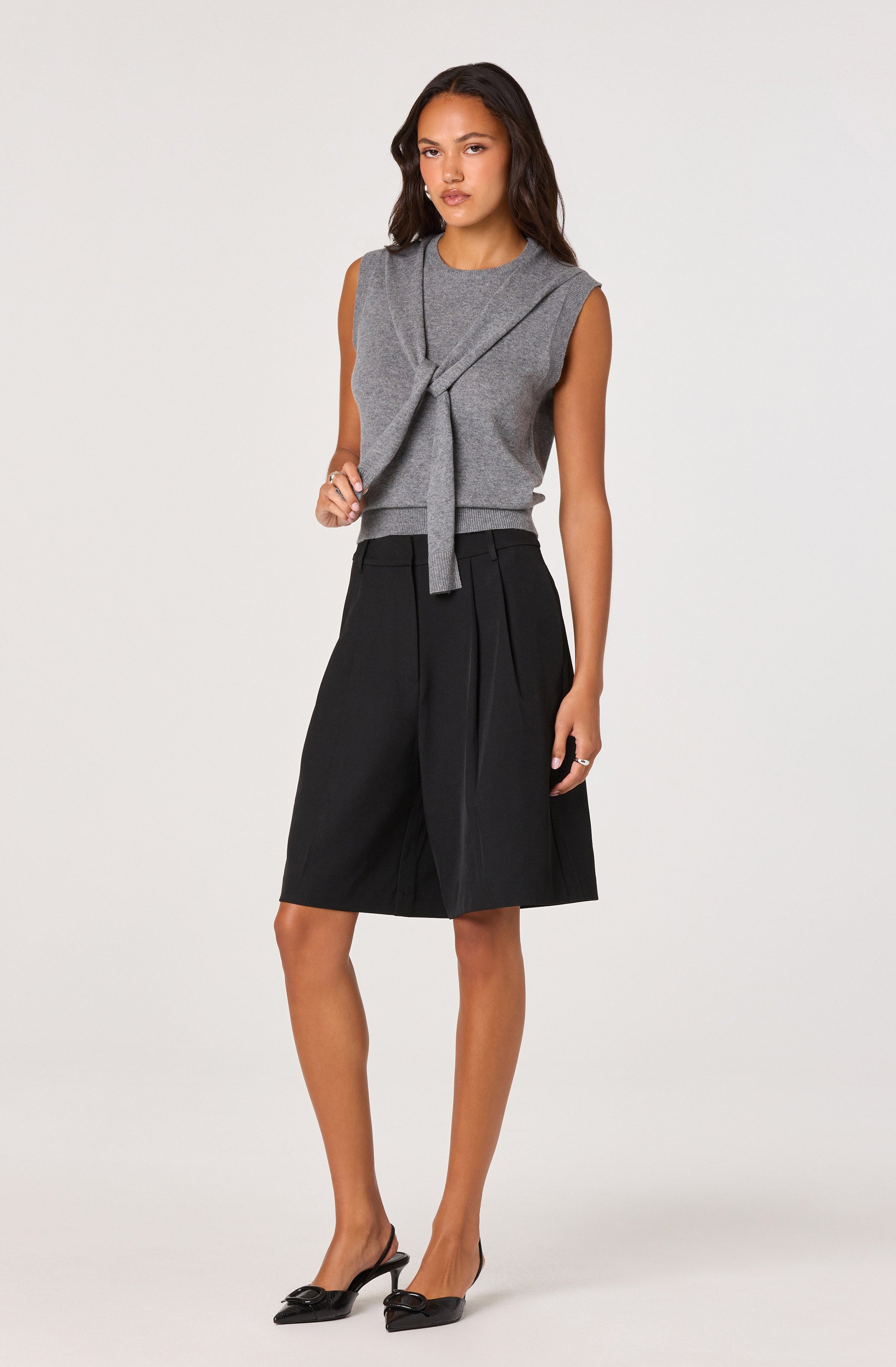 A woman is wearing a sleeveless gray top with a tied detail at the front, paired with a high-waisted, knee-length black skirt featuring pleats. She is also wearing black pointed-toe heels with bows on the front.