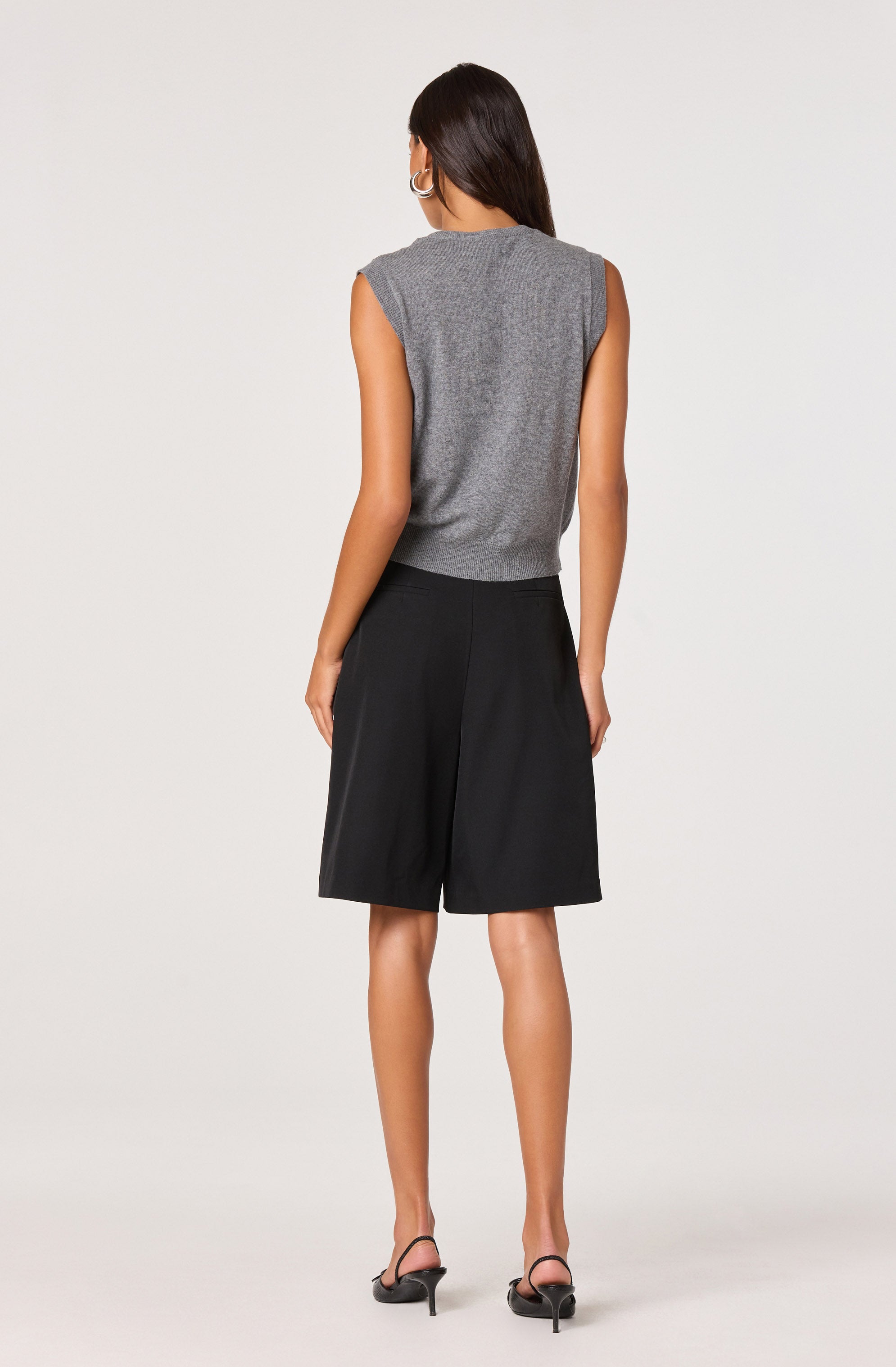 A woman wearing a sleeveless, gray knit top with shoulder pads and black knee-length wide-leg shorts. She is also wearing black strappy high-heeled sandals and hoop earrings.