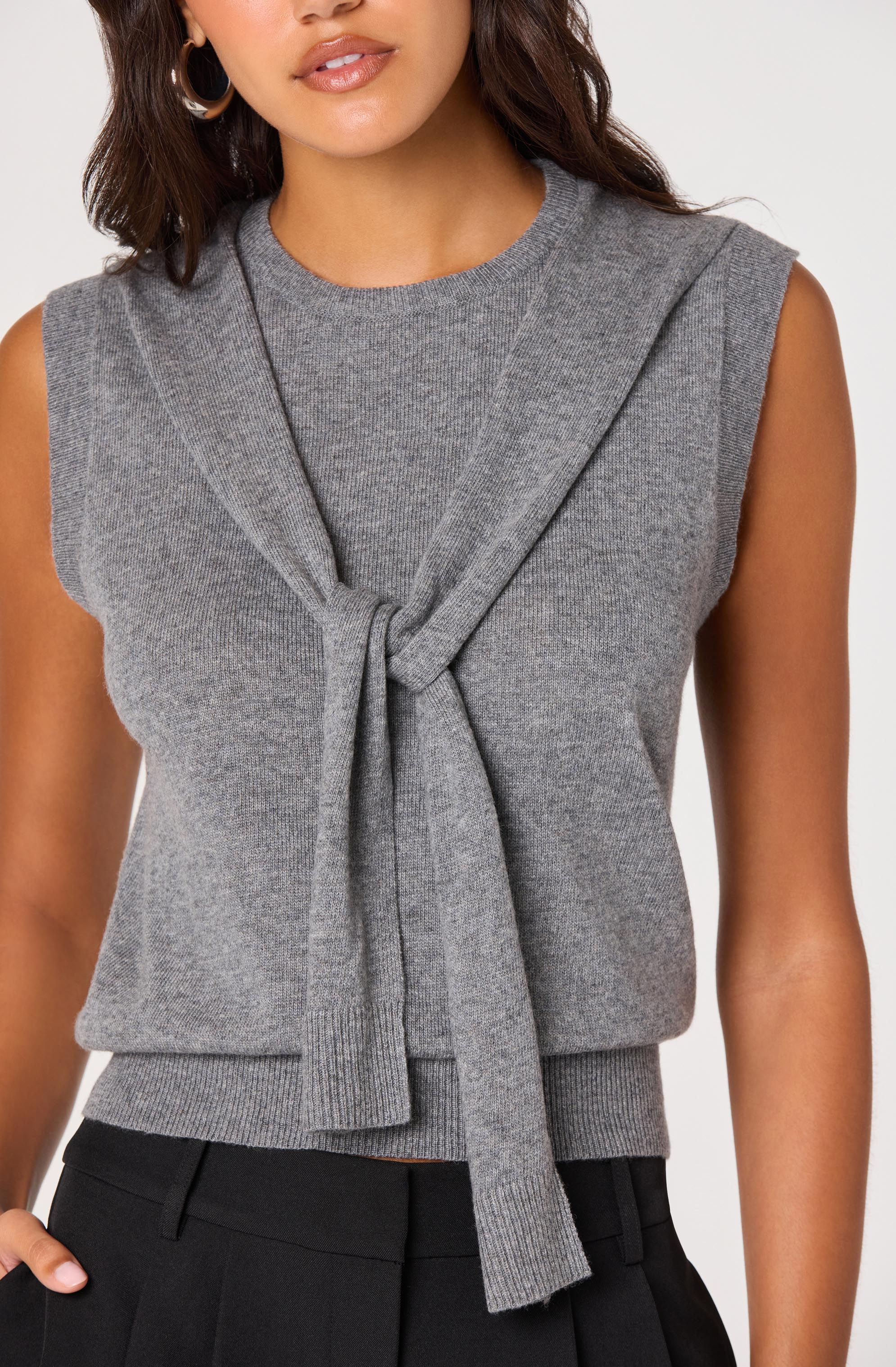 A woman is wearing a sleeveless, grey knit top with a round neckline. The top features a front knot detail with a matching grey knitted tie or scarf draped over her shoulders. She is also wearing black pants.