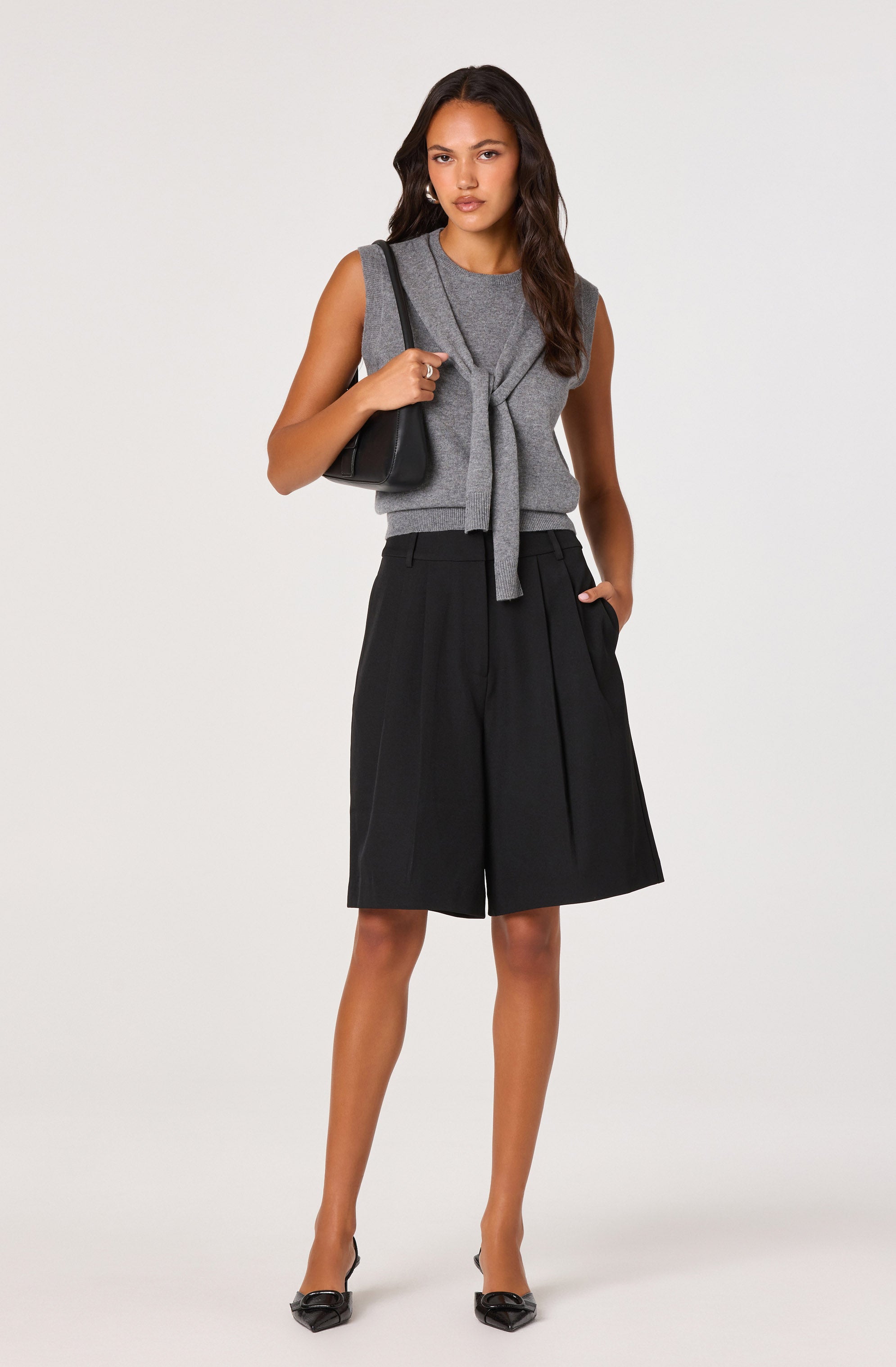 A woman is dressed in a sleeveless gray top with a tied knot at the front, paired with black wide-leg shorts that have pleats. She carries a black shoulder bag and wears pointed black heels.