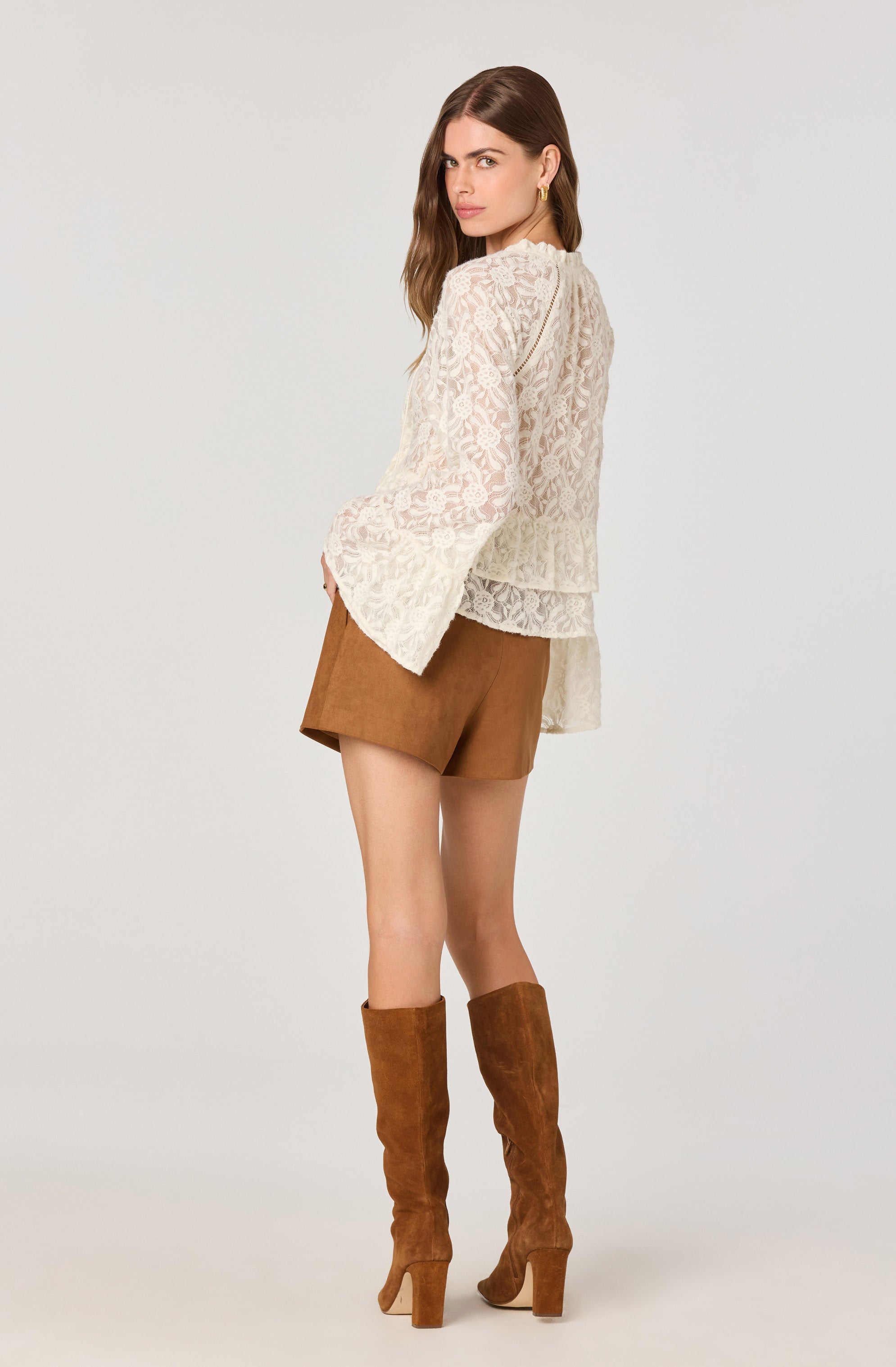 A woman is wearing a long-sleeved, cream-colored lace top with a floral pattern and bell sleeves, paired with high-waisted brown shorts and knee-high brown suede boots with block heels.
