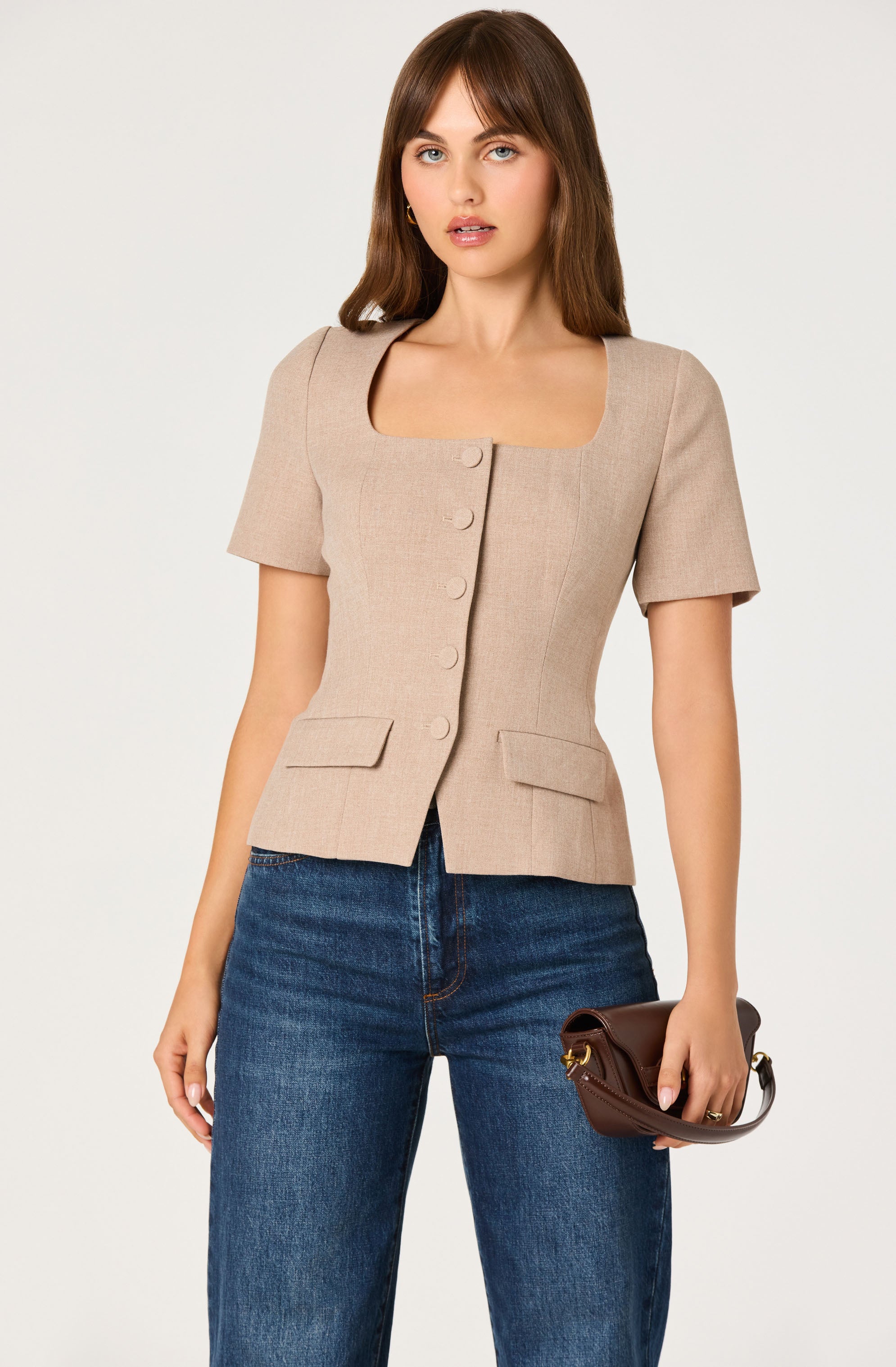 A woman is wearing a beige, short-sleeved blazer with large buttons down the front, two flap pockets at the waist, and a square neckline. She is also holding a small brown handbag with a gold clasp. She has medium-length brown hair and is standing against a plain, light background.