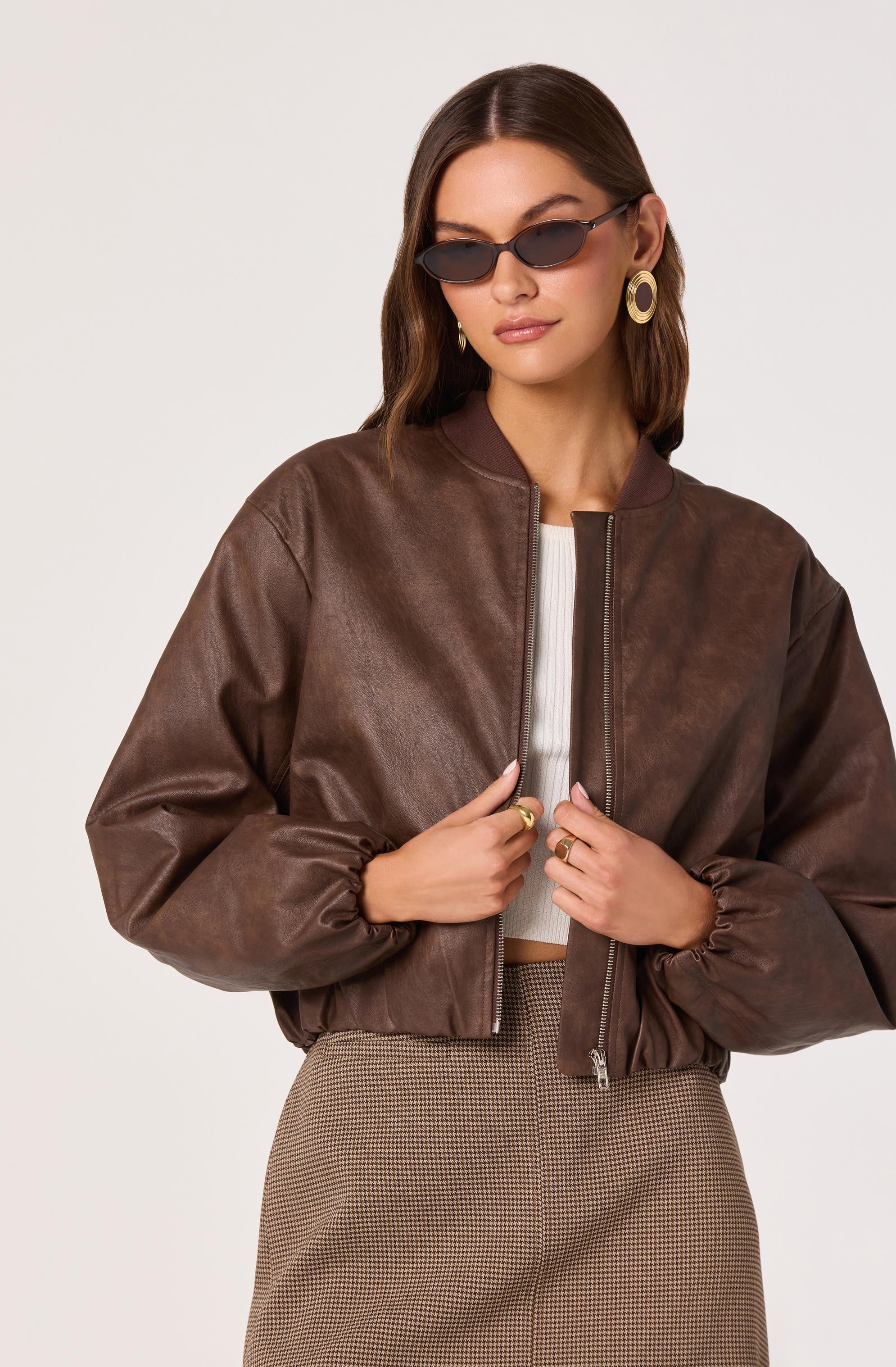 A woman is wearing a brown leather bomber jacket with a front zipper, a white ribbed crop top underneath, and high-waisted checkered pants. She has on large, round earrings, dark sunglasses, and a ring on each hand.