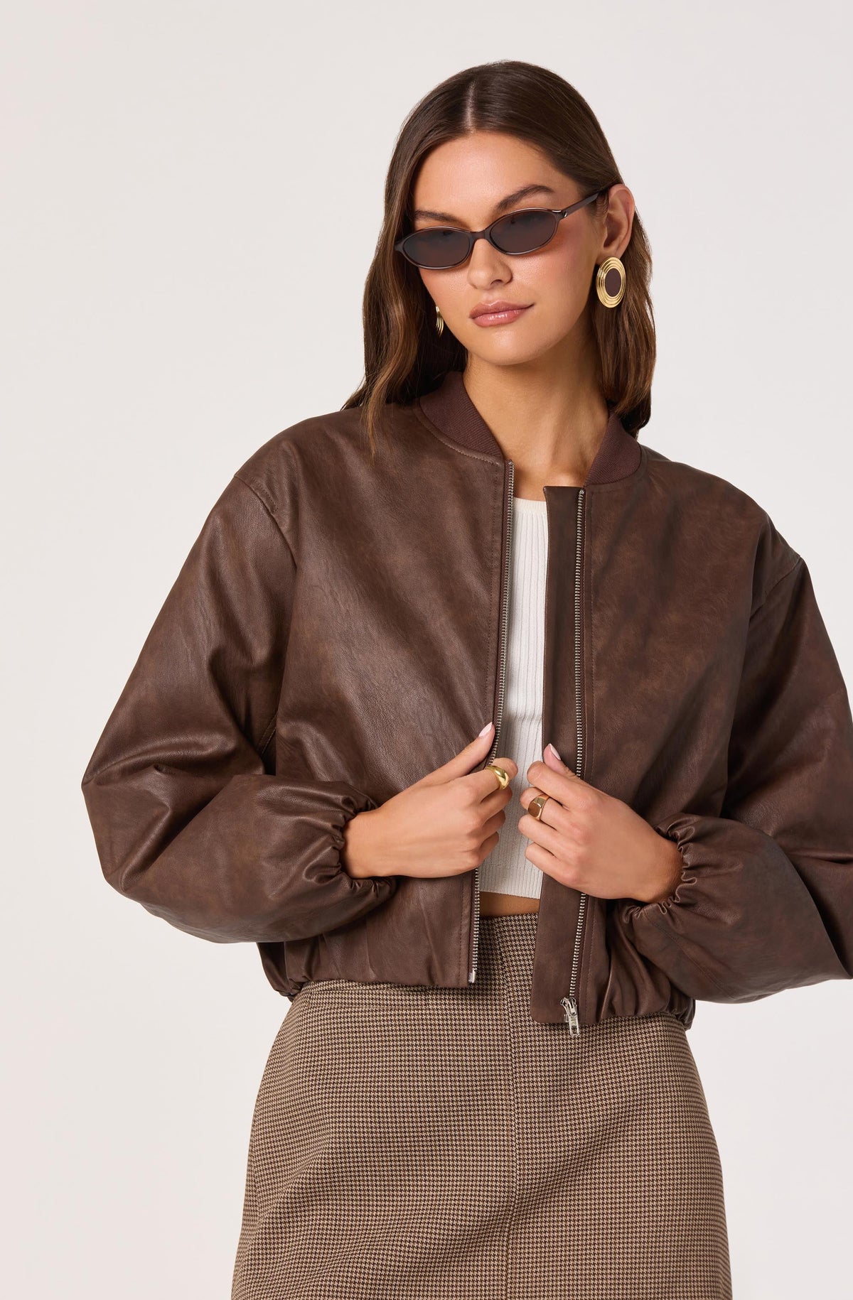 A woman is wearing a brown leather bomber jacket with a front zipper, a white ribbed crop top underneath, and high-waisted checkered pants. She has on large, round earrings, dark sunglasses, and a ring on each hand.