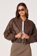 A woman is wearing a brown leather bomber jacket with a front zipper, a white ribbed crop top underneath, and high-waisted checkered pants. She has on large, round earrings, dark sunglasses, and a ring on each hand.