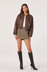 A woman is wearing a brown leather bomber jacket with a zipper front and puffed long sleeves, paired with a white ribbed crop top and a high-waisted, short, brown checkered skirt. She has accessorized with large circular earrings and is wearing tall black knee-high boots with high heels.
