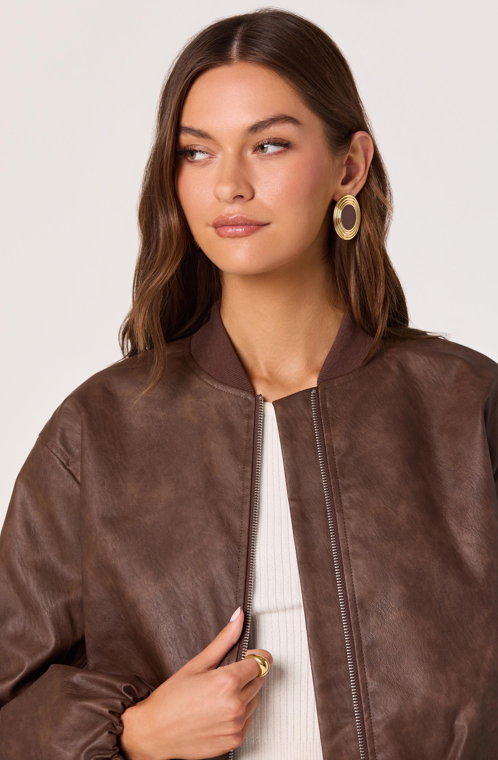 A woman is wearing a brown leather bomber jacket with a ribbed collar, paired with a white ribbed top underneath. She has large circular gold earrings and is also wearing a gold ring on her finger.