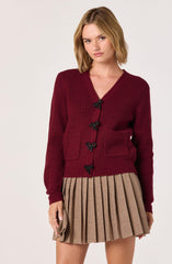 A young woman is wearing a burgundy knit cardigan with black bow-shaped buttons and two front pockets. She is also wearing a tan plaid pleated skirt with thin black and red lines.