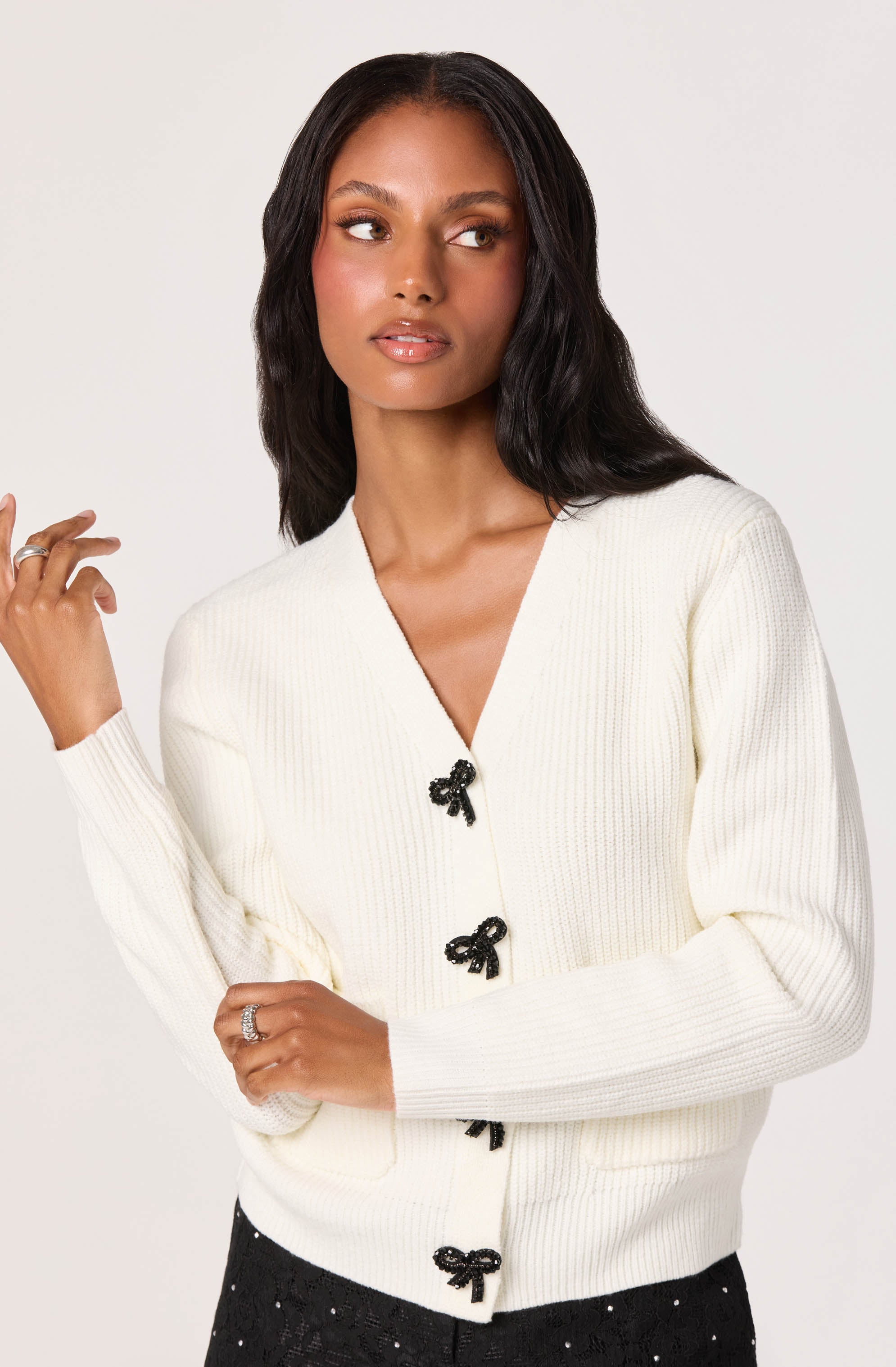 A woman wearing a white knitted cardigan with black bow-shaped buttons down the front. The cardigan has a V-neckline and long sleeves.