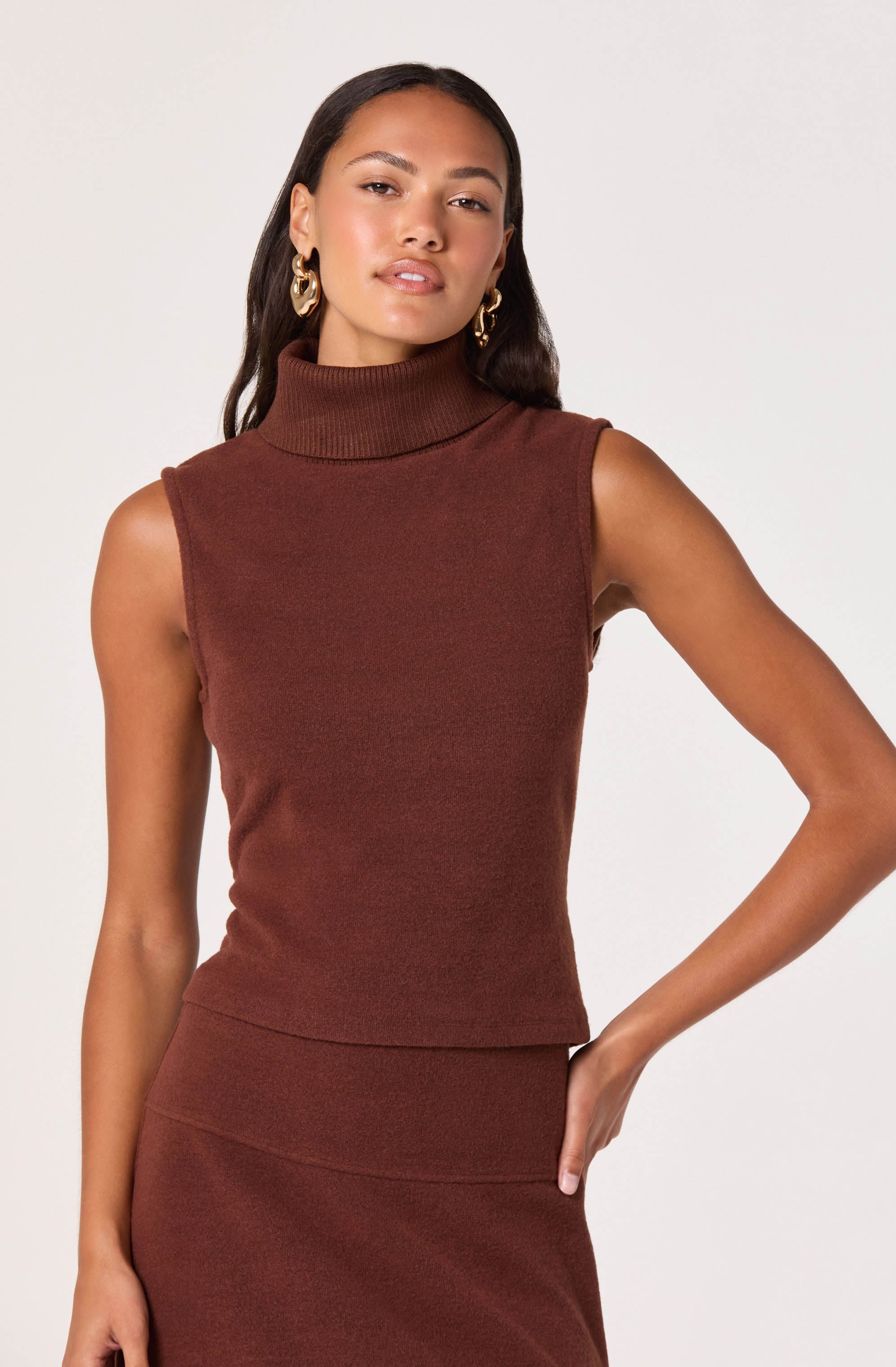 A woman wearing a sleeveless, high-neck, rust-colored knit top paired with a matching skirt. She has long, dark hair, gold hoop earrings, and a confident expression.
