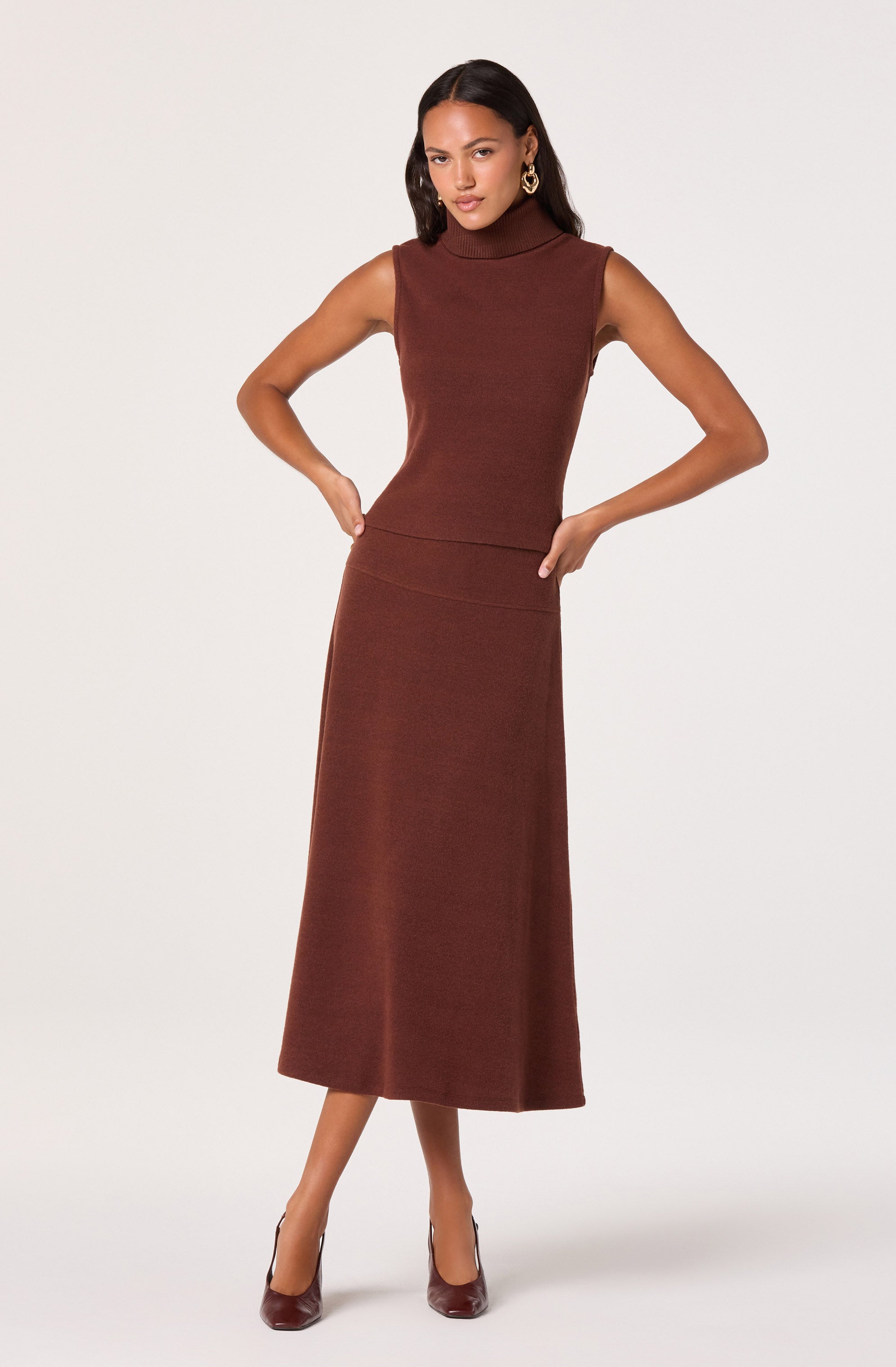 A woman is wearing a sleeveless, high-neck top in a rich brown color, paired with a matching long, fitted skirt that reaches her ankles. She has on matching brown high heels and gold hoop earrings.