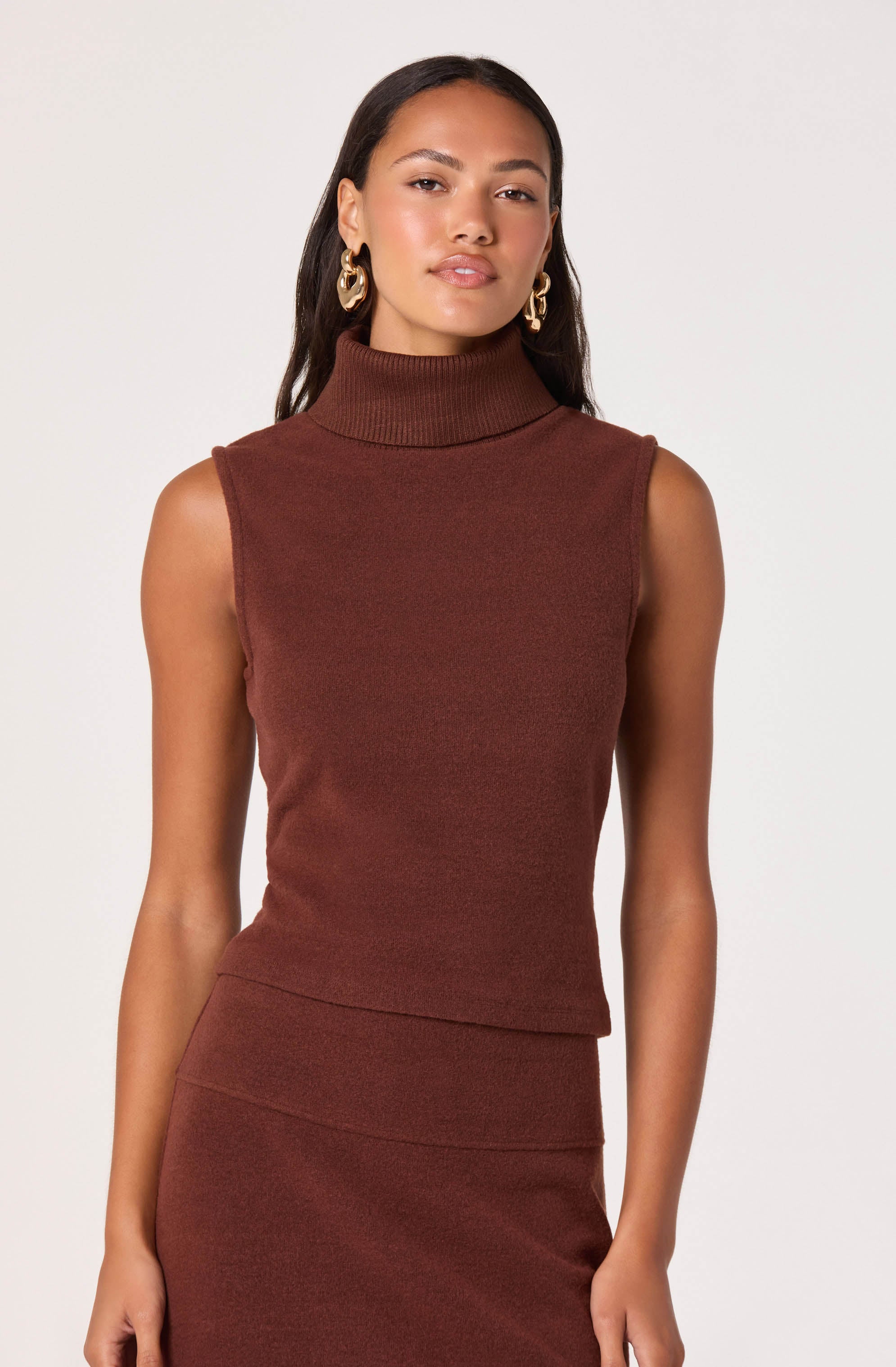 A woman is wearing a sleeveless, high-neck, ribbed knit top in a deep brown color. She has matching brown knit skirt that sits high on her waist, creating a coordinated look. The outfit is accessorized with large, gold hoop earrings.