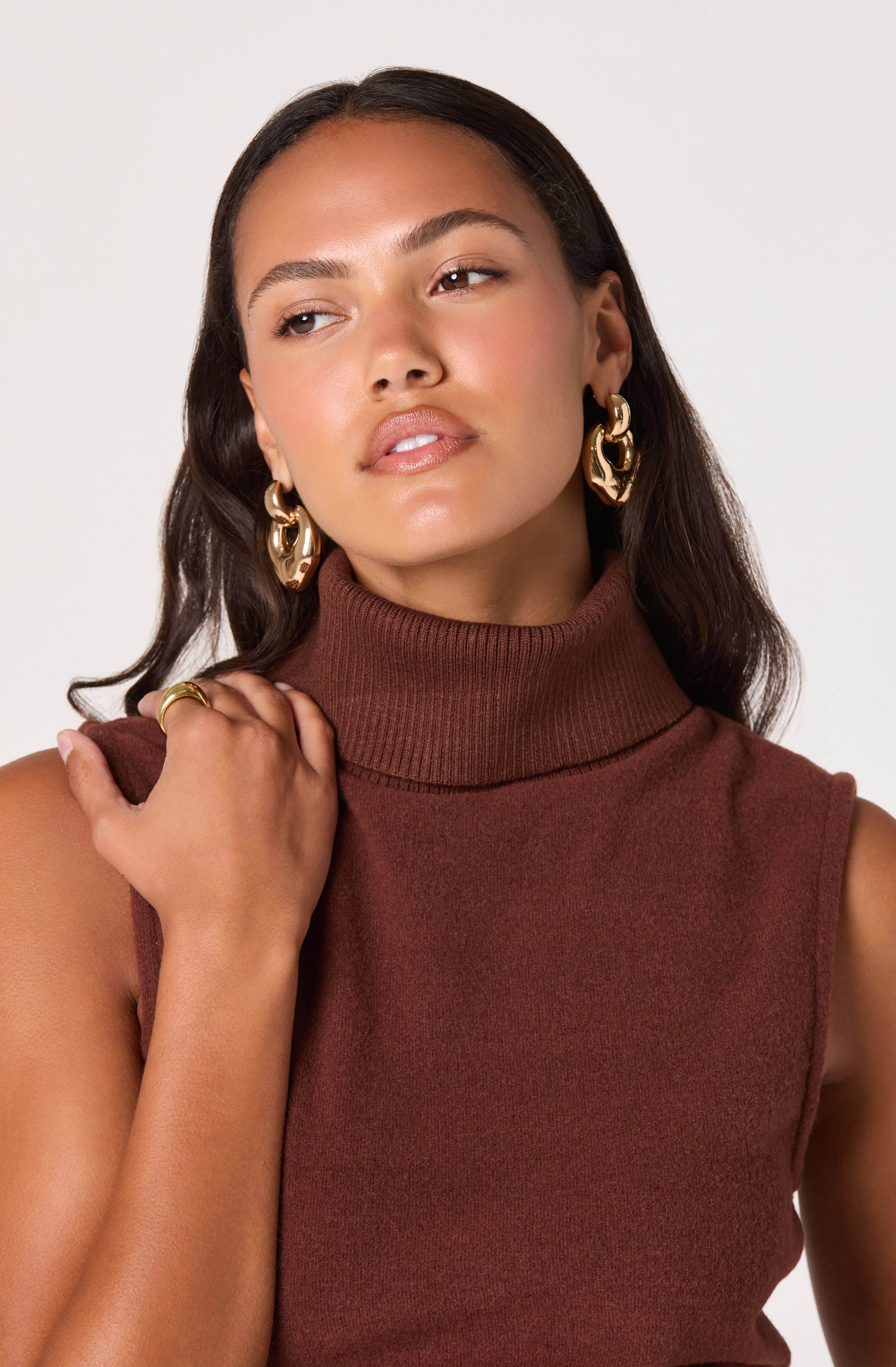A woman with long, dark brown hair is wearing a sleeveless, high-neck, brick-red ribbed top. She has large, gold statement hoop earrings and a matching gold ring on her finger.