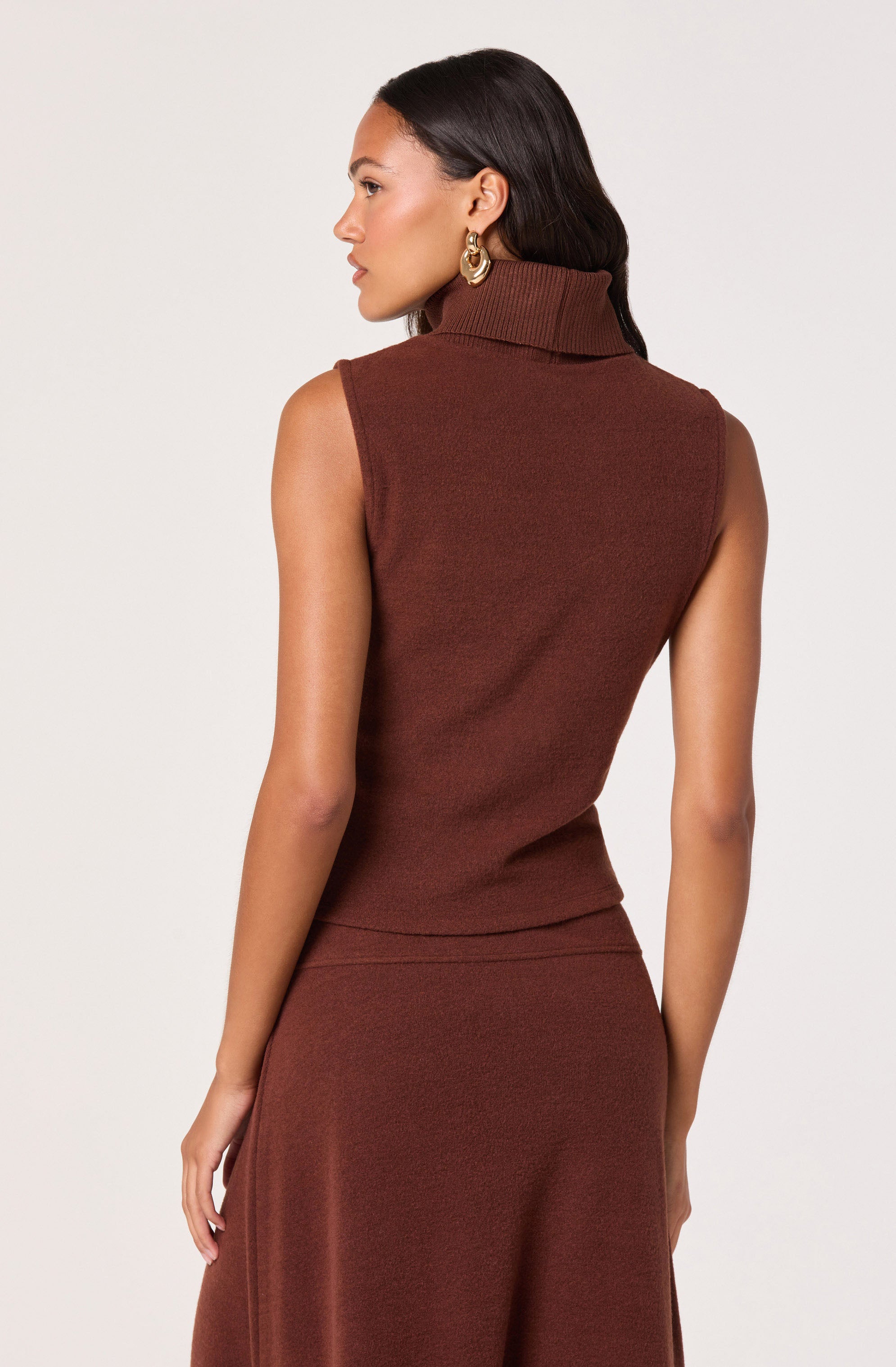 A woman is wearing a sleeveless, fitted, rust-colored turtleneck top with a ribbed collar, paired with a matching high-waisted skirt. She has long, dark hair and is accessorizing with large gold hoop earrings.