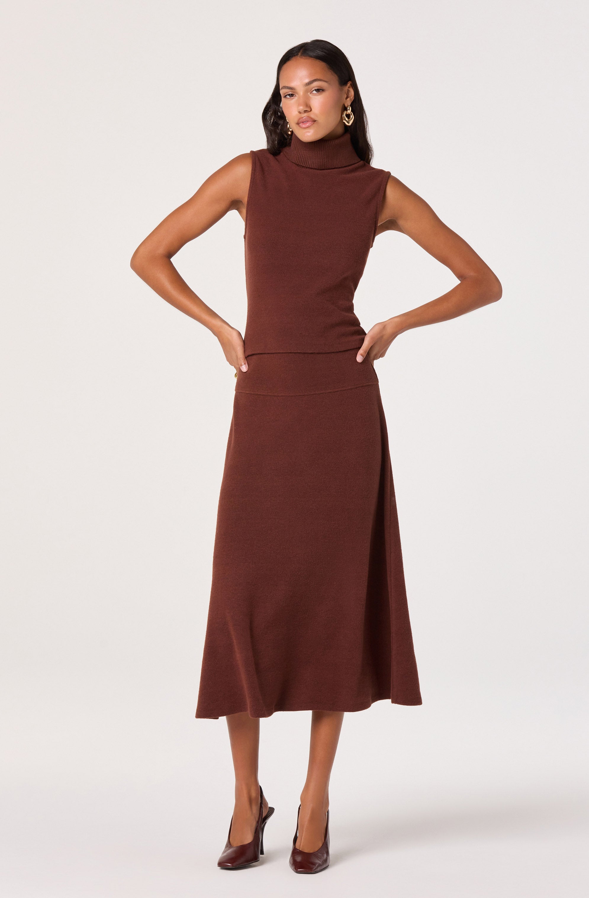 A woman is wearing a sleeveless, high-neck, rust-colored knit top paired with a matching long, A-line skirt that has a side slit. She is also wearing dark brown high-heeled shoes and gold earrings.