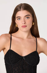 A woman is wearing a black lace camisole with thin spaghetti straps. The camisole features delicate lace fabric with floral patterns and a small bow detail at the center of the bust.