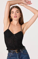 A woman wearing a black lace camisole top with thin spaghetti straps and a scalloped neckline. The top has a fitted style with a subtle floral lace pattern and a slight peplum hem. She is also wearing high-waisted blue jeans with a black belt featuring a gold heart-shaped buckle.