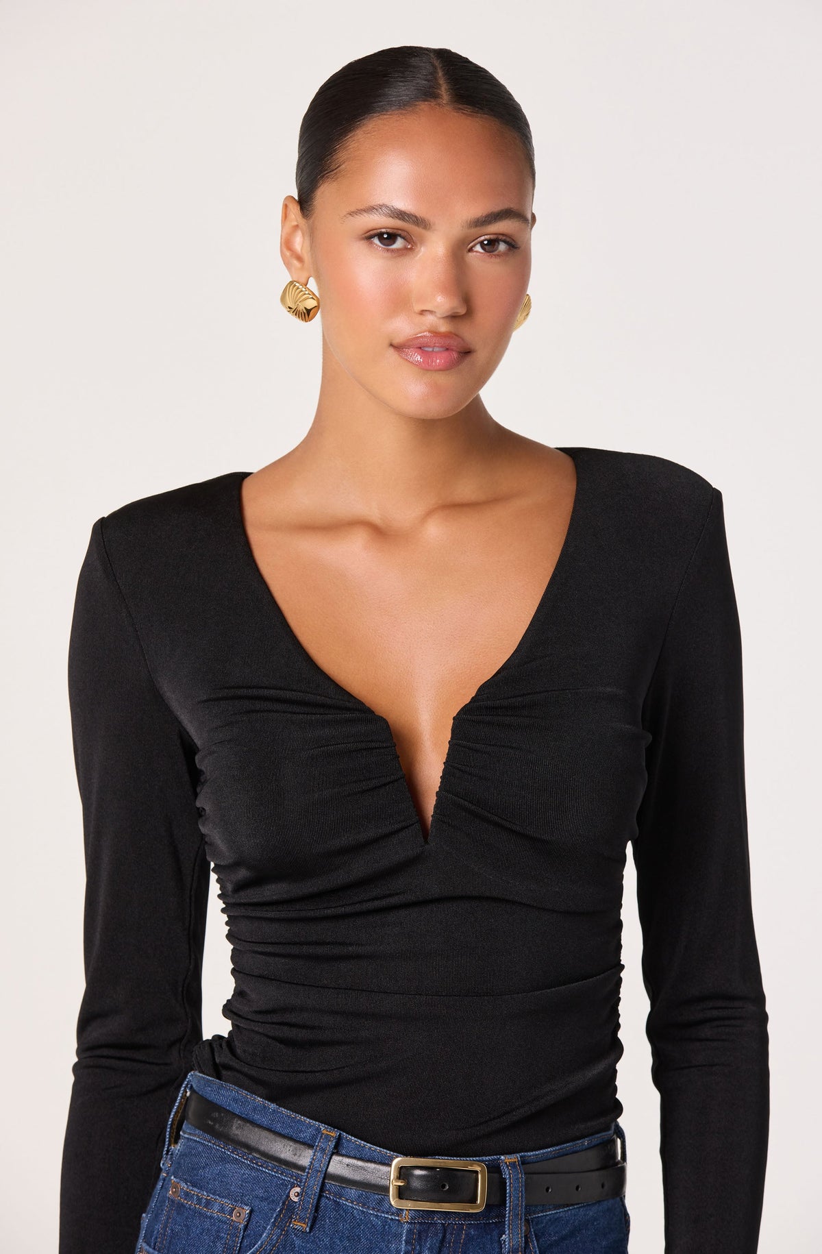 A woman is wearing a black long-sleeve top with a deep V neckline and ruched detailing at the front. She has on gold statement earrings and dark blue jeans with a black belt featuring a gold buckle.
