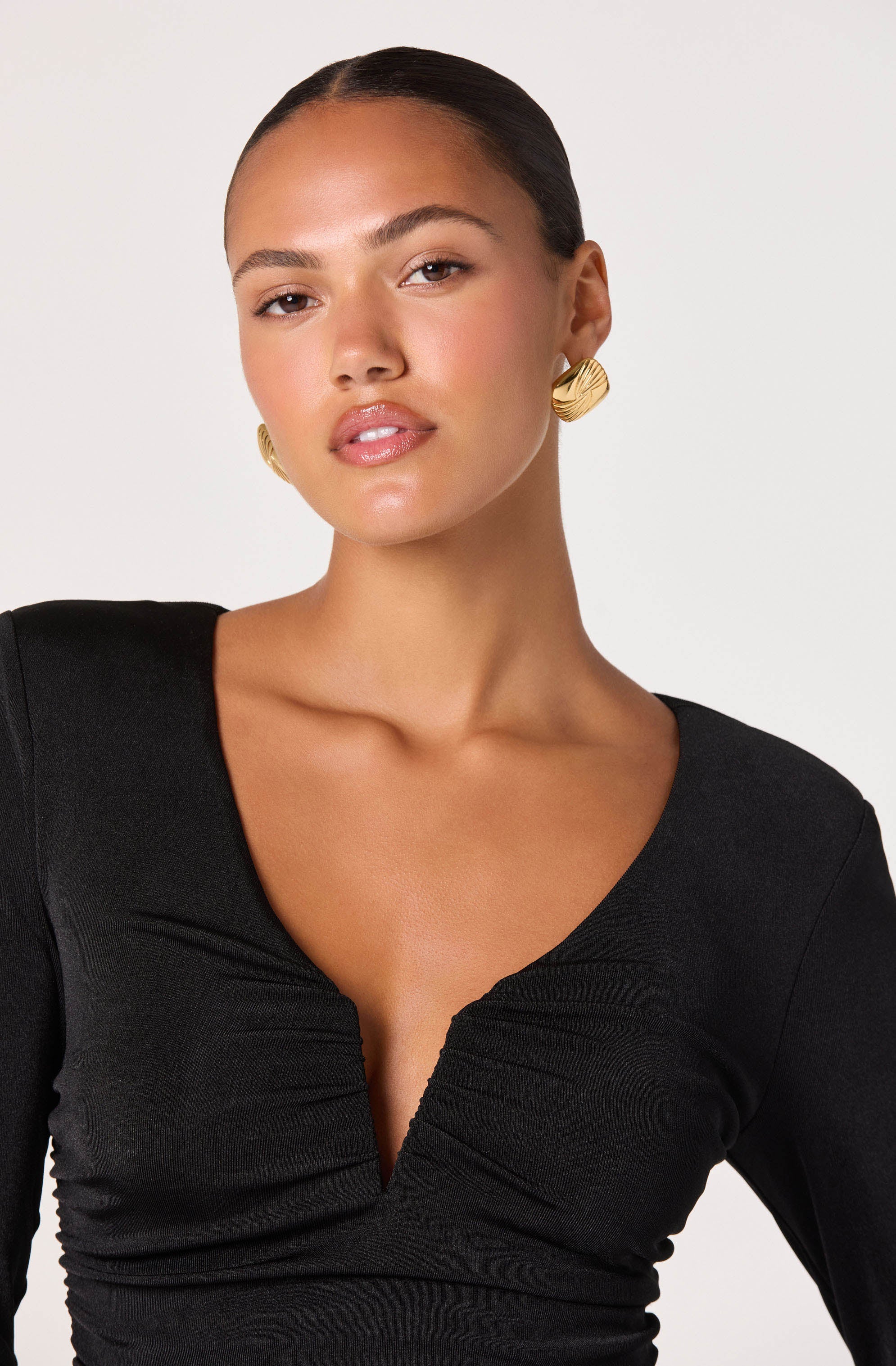 A woman is wearing a fitted black top with a deep V neckline that features gathered detailing at the center. She has on gold, textured, circular earrings.