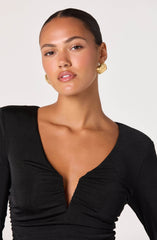 A woman is wearing a fitted black top with a deep V neckline that features gathered detailing at the center. She has on gold, textured, circular earrings.