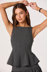 A woman is wearing a sleeveless, fitted, gray dress with a subtle checkered pattern. The dress features a round neckline and princess seams that create a tailored fit. It has a flared, ruffled peplum at the waist that extends to a pleated skirt with a ruffled hem. She accessorizes with large gold hoop earrings and a black bracelet on her right wrist. The woman has shoulder-length, wavy brown hair and is posing with her arms raised above her head against a plain, light background.