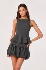A young woman with long brown hair is wearing a sleeveless, dark gray patterned top with a peplum waist and matching voluminous shorts that are gathered at the hem. She has minimal makeup and is accessorized with gold earrings and a black bracelet.