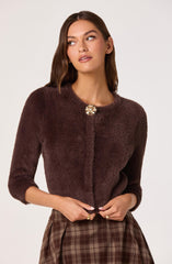 A woman is wearing a dark brown, fuzzy, long-sleeved jacket with a round neckline, fastened at the top with a decorative brooch that resembles a flower. She is also wearing a brown plaid skirt.