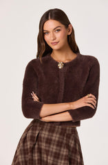 A woman is wearing a dark brown, fuzzy, cropped cardigan with three-quarter length sleeves and a decorative brooch at the neckline. She is also dressed in a high-waisted, knee-length skirt featuring a brown plaid pattern.