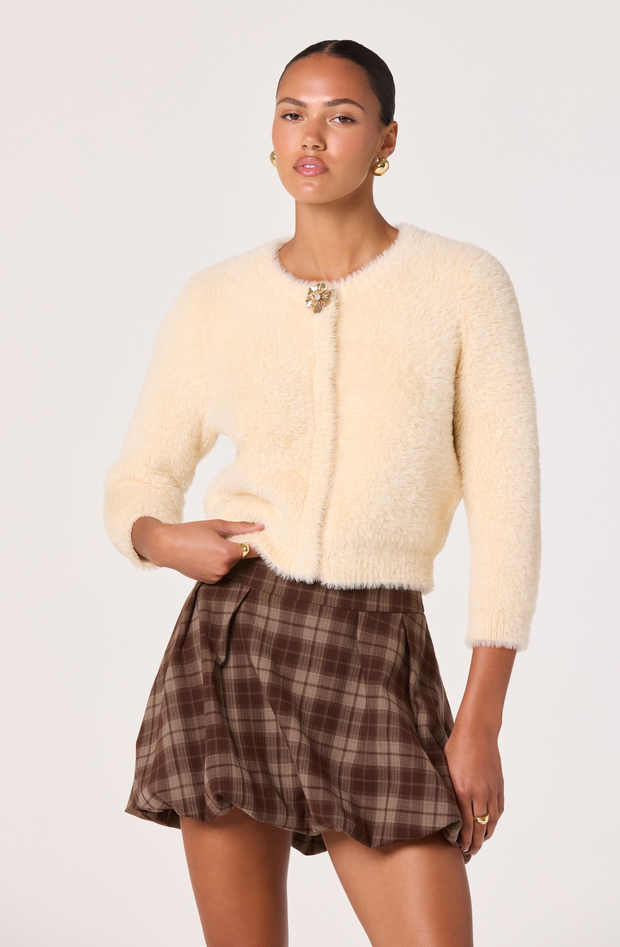 A woman is wearing a soft, cream-colored, fuzzy cardigan with three-quarter sleeves and a decorative brooch at the neckline. She is also wearing high-waisted, loose-fitting brown plaid shorts with pleats. She has gold hoop earrings and a gold ring on her right hand.