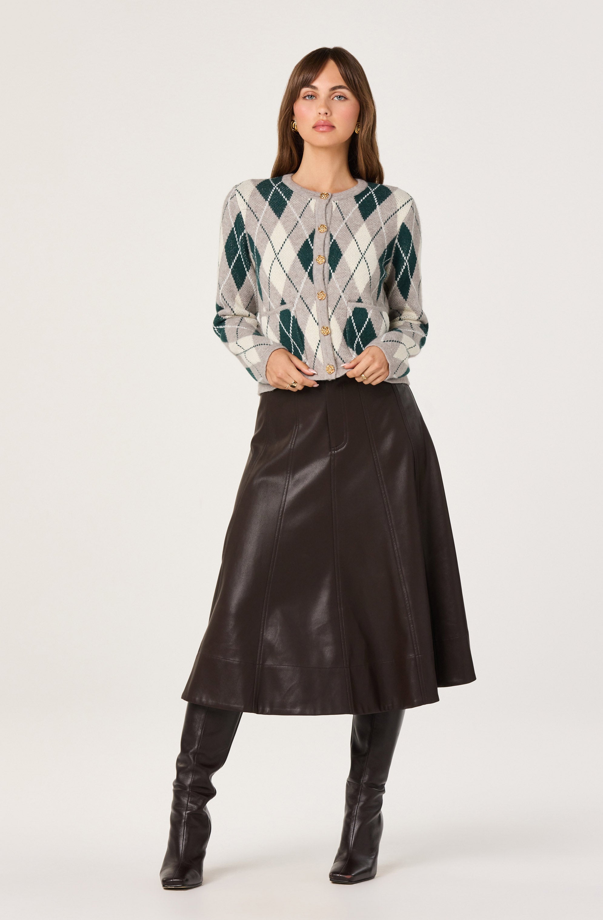The woman is wearing a light-colored button-up cardigan with an argyle pattern in shades of green, beige, and white. She pairs it with a dark brown, leather A-line skirt that falls below the knees. She completes the look with tall, dark leather boots that reach just above the knees.