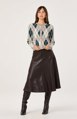 The woman is wearing a light-colored button-up cardigan with an argyle pattern in shades of green, beige, and white. She pairs it with a dark brown, leather A-line skirt that falls below the knees. She completes the look with tall, dark leather boots that reach just above the knees.