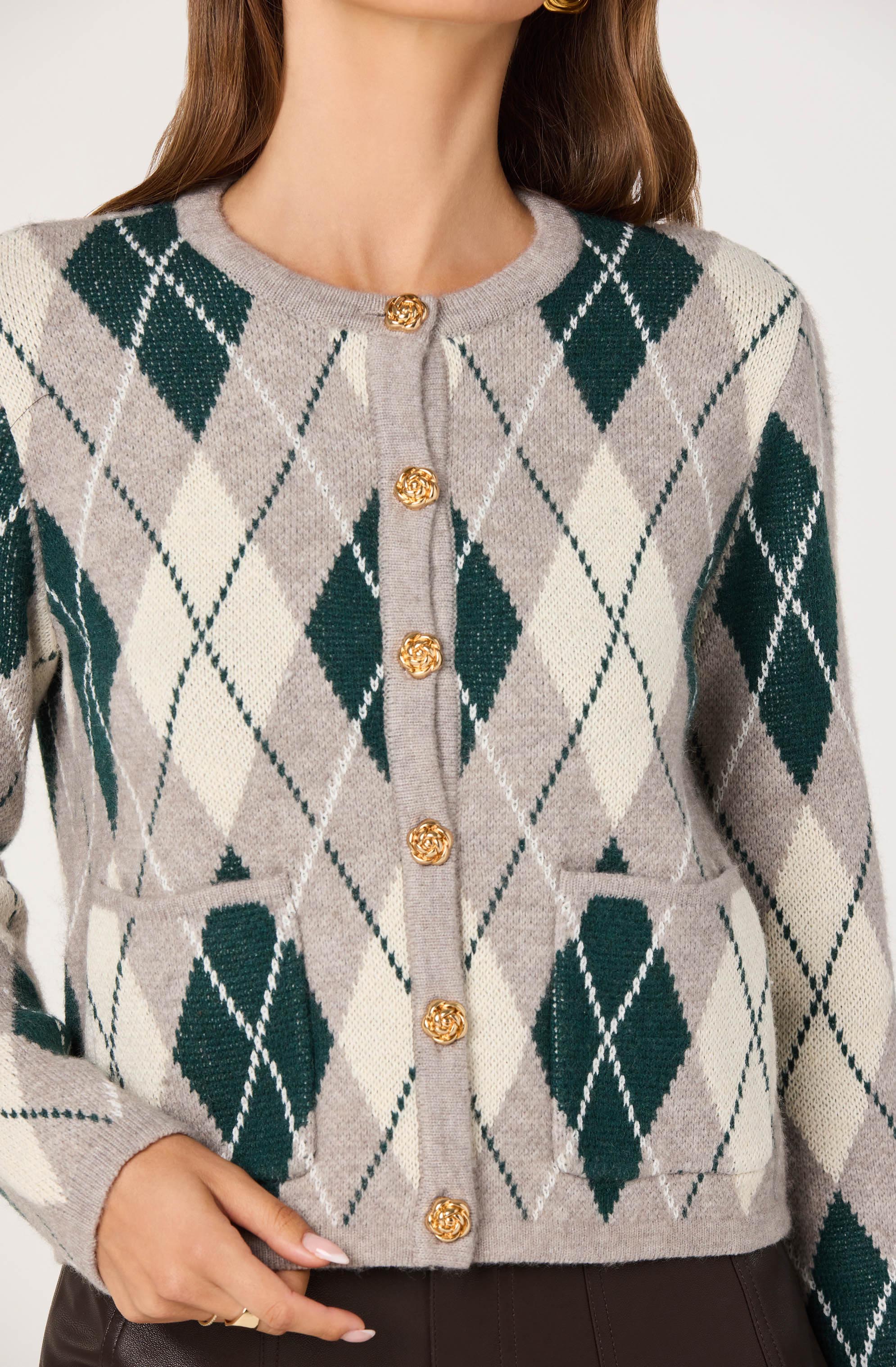 A woman is wearing a cardigan with a classic argyle pattern. The cardigan features a light gray base color with large, dark green diamonds and smaller, cream-colored diamonds intersected by thin, dark green lines. It has a round neckline and is fastened down the front with gold, floral-shaped buttons. The cardigan also has two front pockets near the waist, aligned with the diamond pattern.