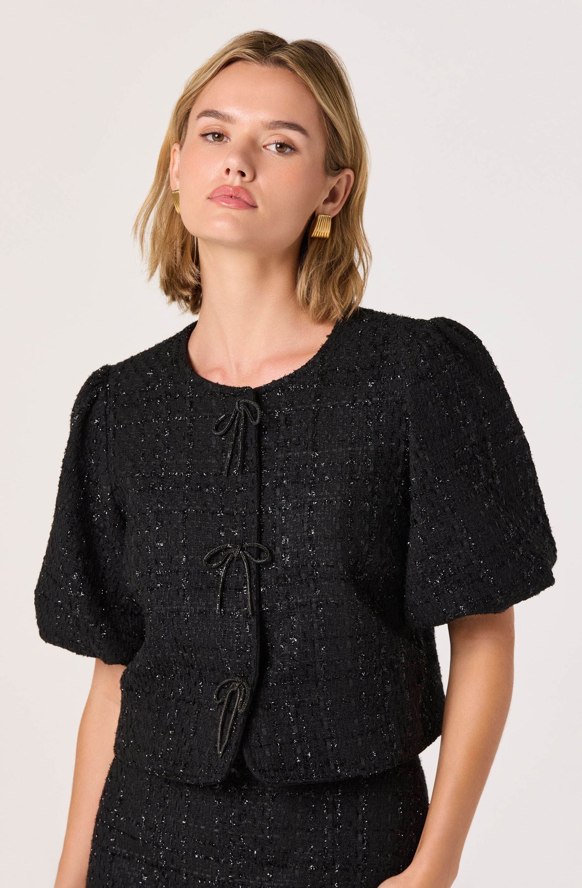 A woman is wearing a black textured short-sleeve jacket with a round neckline and three tie closures down the front. The fabric appears to be a enriched tweed or boucle with a subtle shimmer.