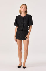 A woman is wearing a black, textured two-piece outfit consisting of a short-sleeved, zip-up jacket with a bow tie at the neckline and a matching high-waisted mini skirt. The fabric has a tweed-like appearance with small multicolored flecks. She completes the look with black pointed-toe flats with a bow detail.