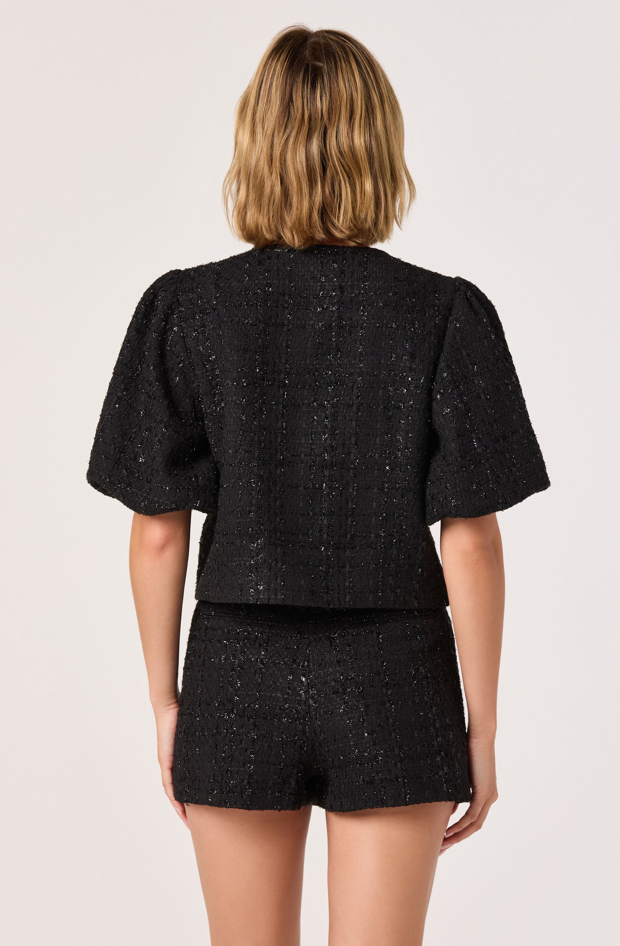 A woman is wearing a black textured two-piece outfit consisting of a short-sleeved top with puff sleeves and matching high-waisted shorts. The fabric appears to have a slightly shiny, speckled finish.