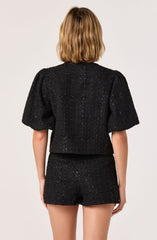 A woman is wearing a black textured two-piece outfit consisting of a short-sleeved top with puff sleeves and matching high-waisted shorts. The fabric appears to have a slightly shiny, speckled finish.
