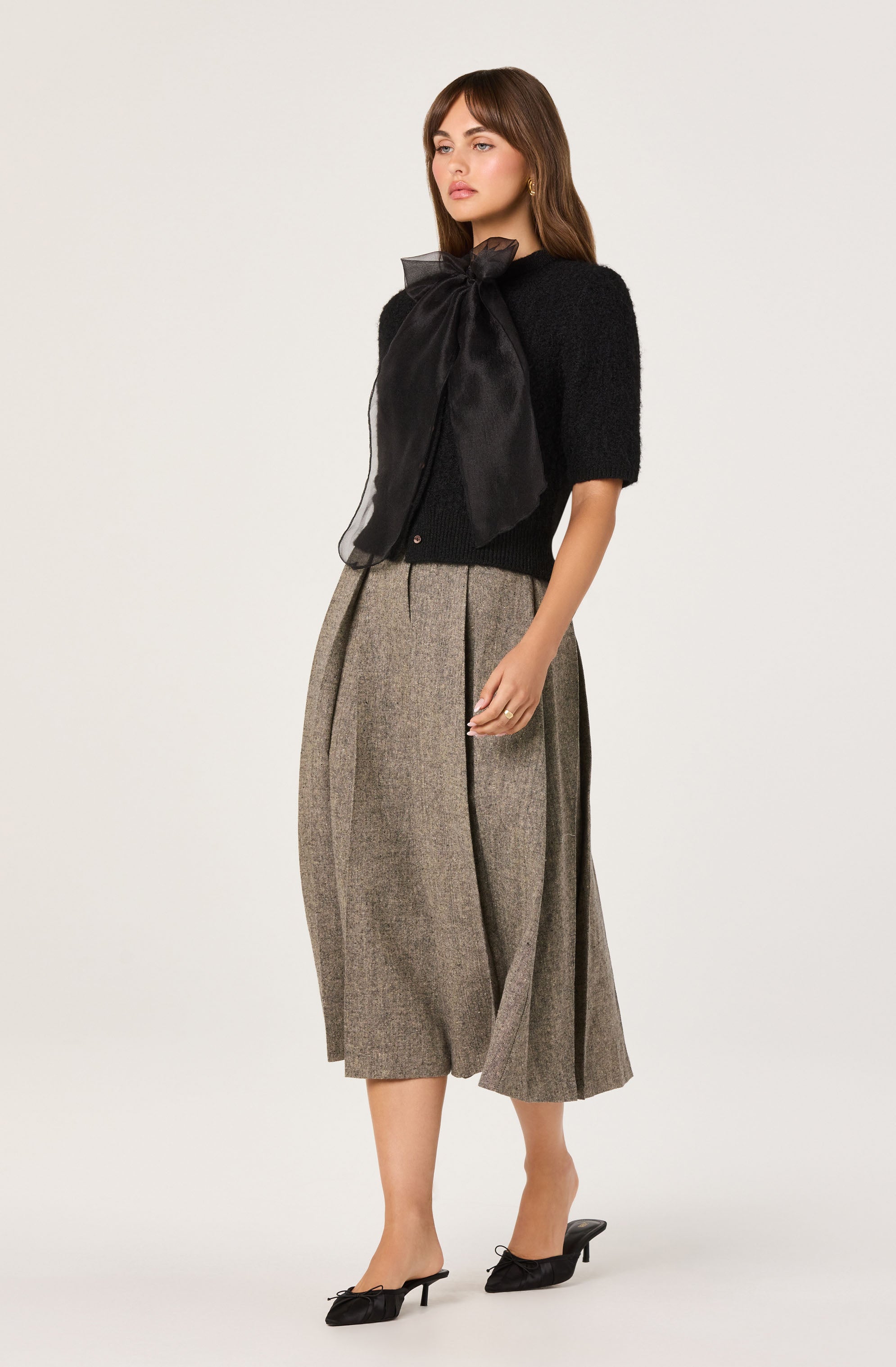 A woman is wearing a black short-sleeved sweater with a large satin bow tied at the neckline. She has on wide-leg, high-waisted, gray plaid culottes that fall to mid-calf, and black pointed-toe mules with a small heel and bow details.