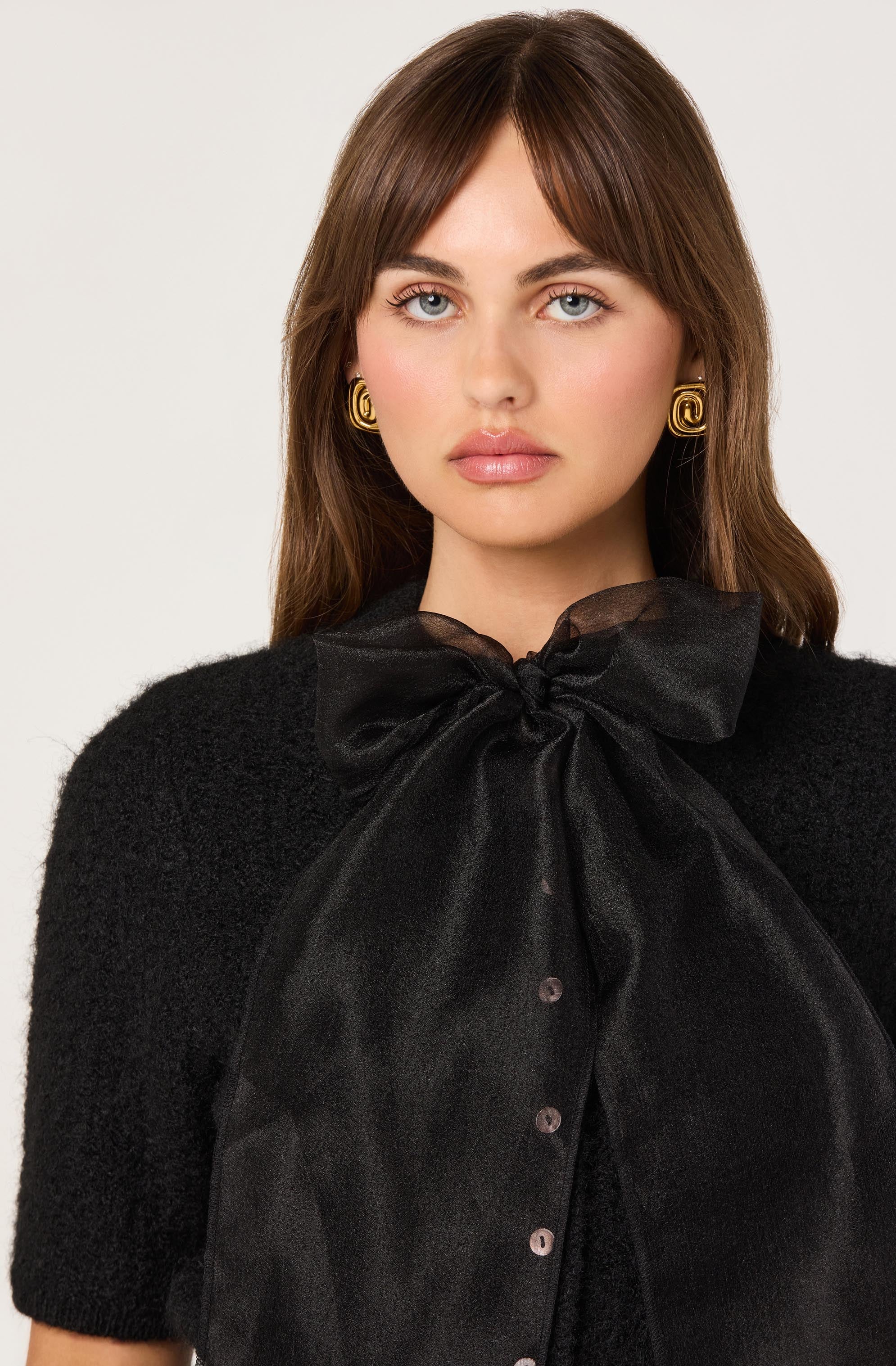 A woman is wearing a black, fuzzy short-sleeve top and a large, sheer black bow tied at the neck with button details down the front. She has gold spiral earrings.