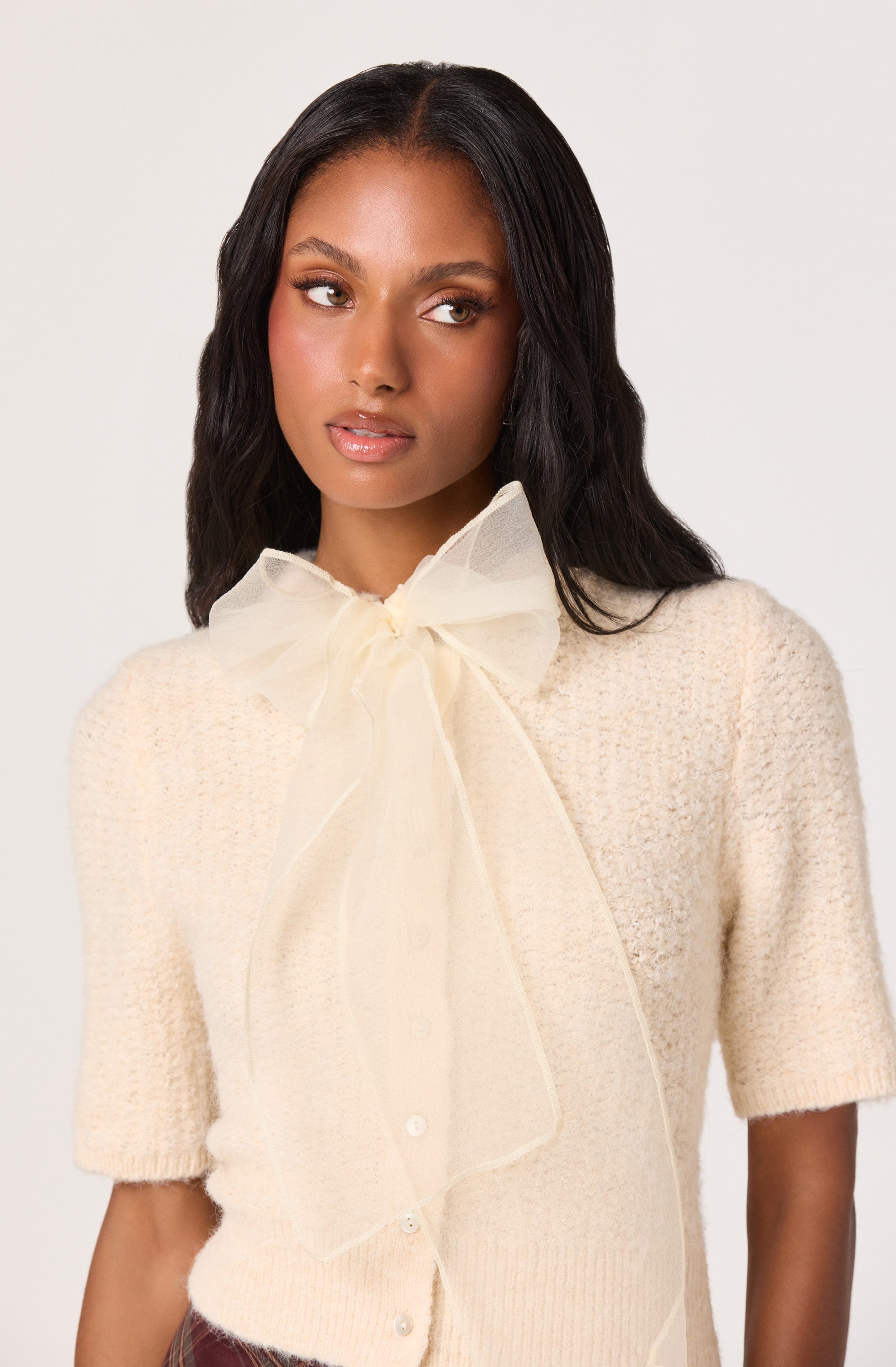 A woman is wearing a cream-colored knitted short-sleeve cardigan with a textured pattern. The cardigan has a button-down front and features a large, sheer, cream-colored bow tied at the collar.