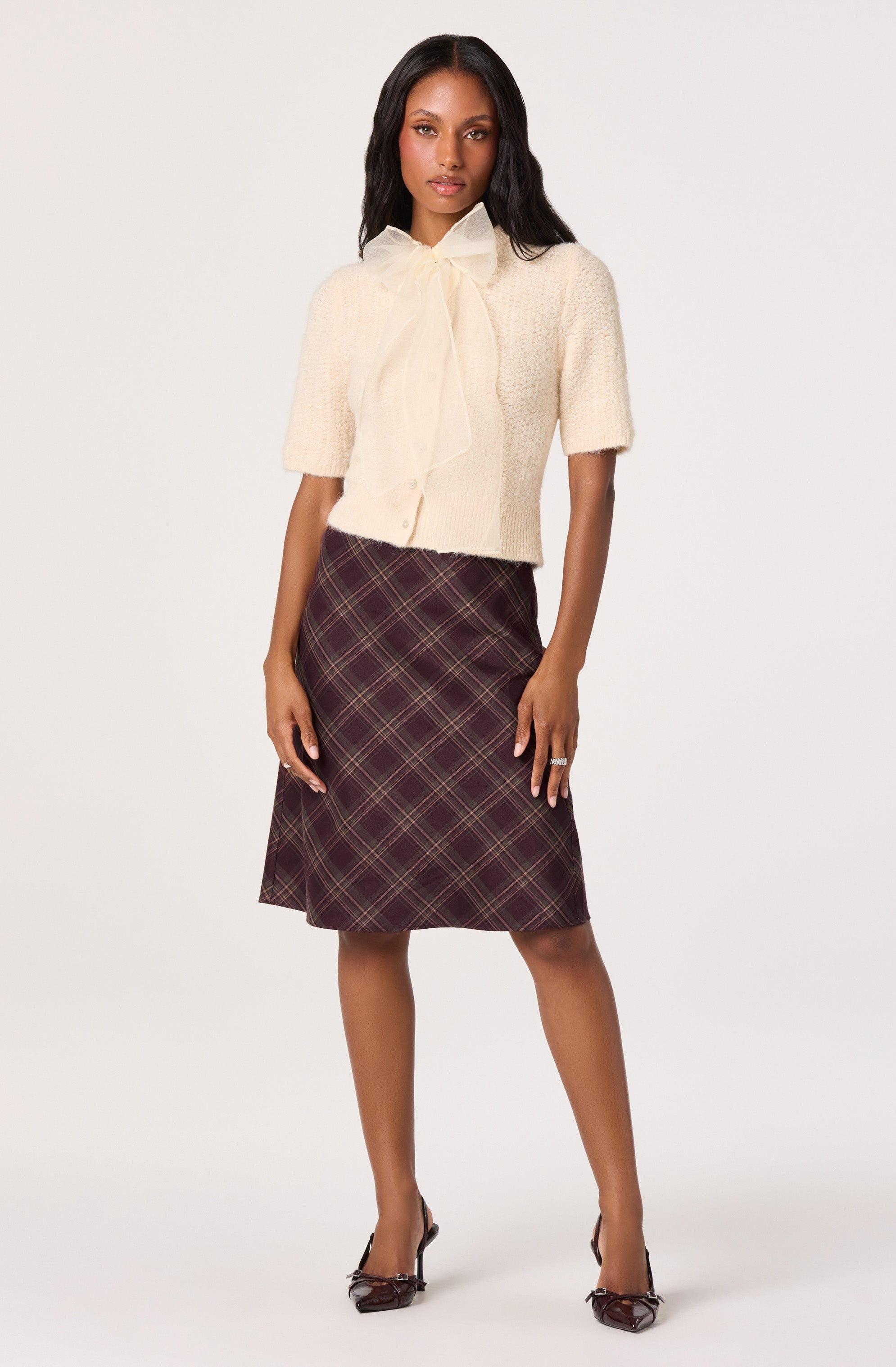 A woman is wearing a cream-colored, short-sleeved cardigan with a large bow tie at the collar made from the same fabric. She pairs it with a knee-length, A-line skirt featuring a dark plaid pattern with red and yellow lines. She completes her outfit with pointed-toe high heels.
