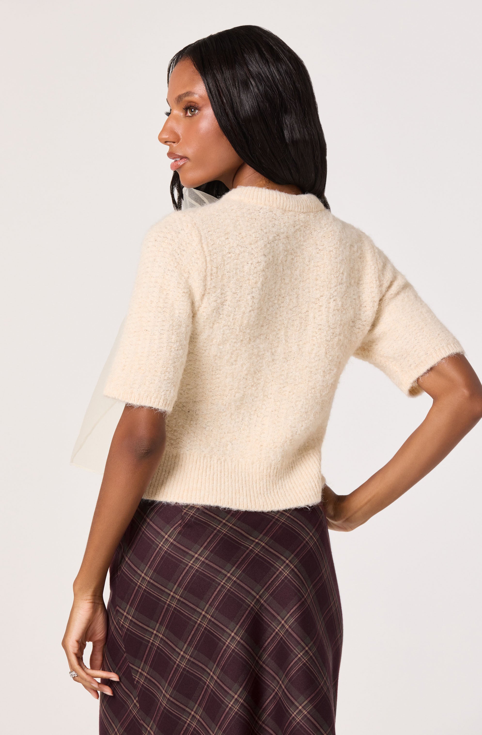 A woman is wearing a cream-colored, short-sleeved, fuzzy knit sweater that has a relaxed fit and a crew neckline. She pairs it with a high-waisted, dark plaid skirt featuring shades of burgundy, green, and beige. The skirt has a classic tartan pattern and a tailored appearance.
