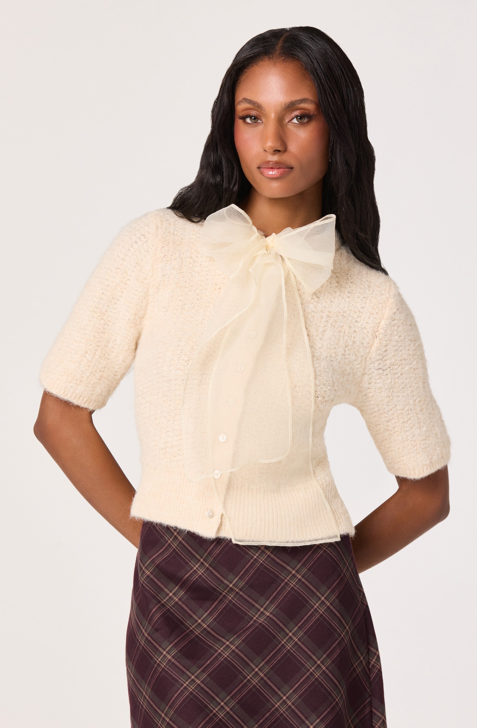 A woman is wearing a cream-colored short-sleeve cardigan with a large, sheer bow tie at the collar. The cardigan is knit with a textured pattern and buttons down the front. She pairs it with a high-waisted, dark plaid skirt featuring intersecting lines in shades of red, green, and yellow.