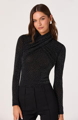 A woman is wearing a long-sleeved, fitted black top with a high, draped neckline. The top is decorated with small, evenly spaced white polka dots throughout. She has paired it with high-waisted black trousers.