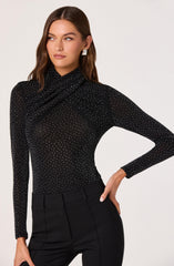 A woman is wearing a fitted, long-sleeved black top adorned with small white dots throughout. The top has a high neckline that drapes slightly, creating a gathered effect around the neck and shoulders. She is also wearing high-waisted black pants with a tailored fit. The woman has minimal makeup and shoulder-length brown hair styled in loose waves. She accessorizes with a pair of large, square, silver earrings.