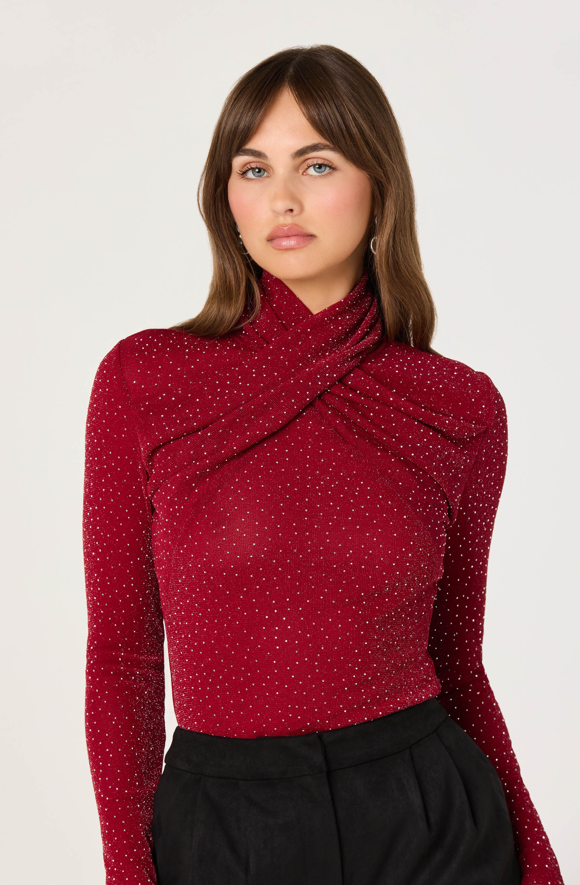 A woman is wearing a fitted, long-sleeve, red top with a high neckline and a twisted, draped design across the chest. The top is decorated with small, evenly spaced white polka dots. She is also wearing black high-waisted pants.