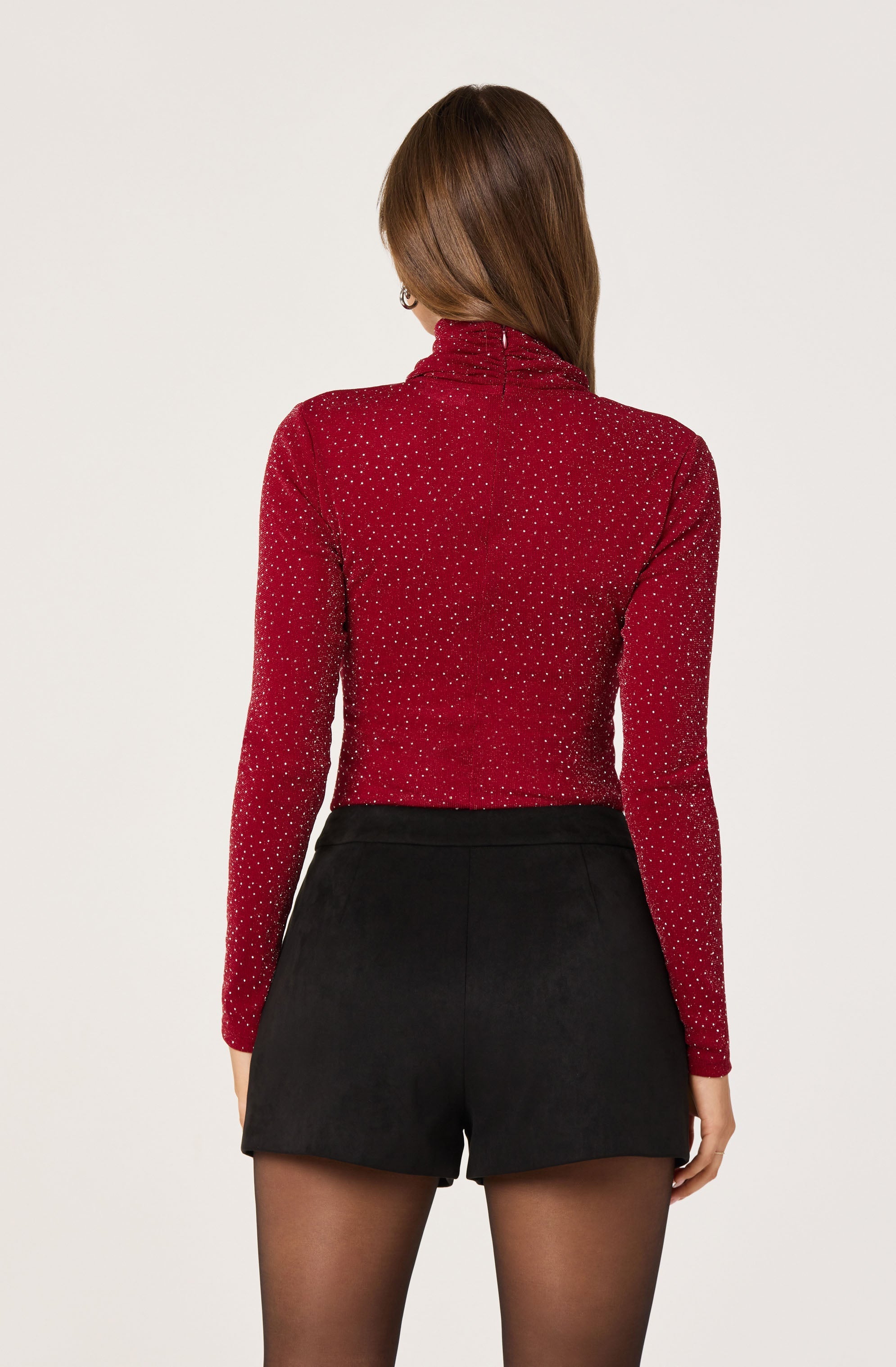 A woman is wearing a fitted, long-sleeve, red turtleneck sweater with small white polka dots and black high-waisted shorts. She has long, straight brown hair.