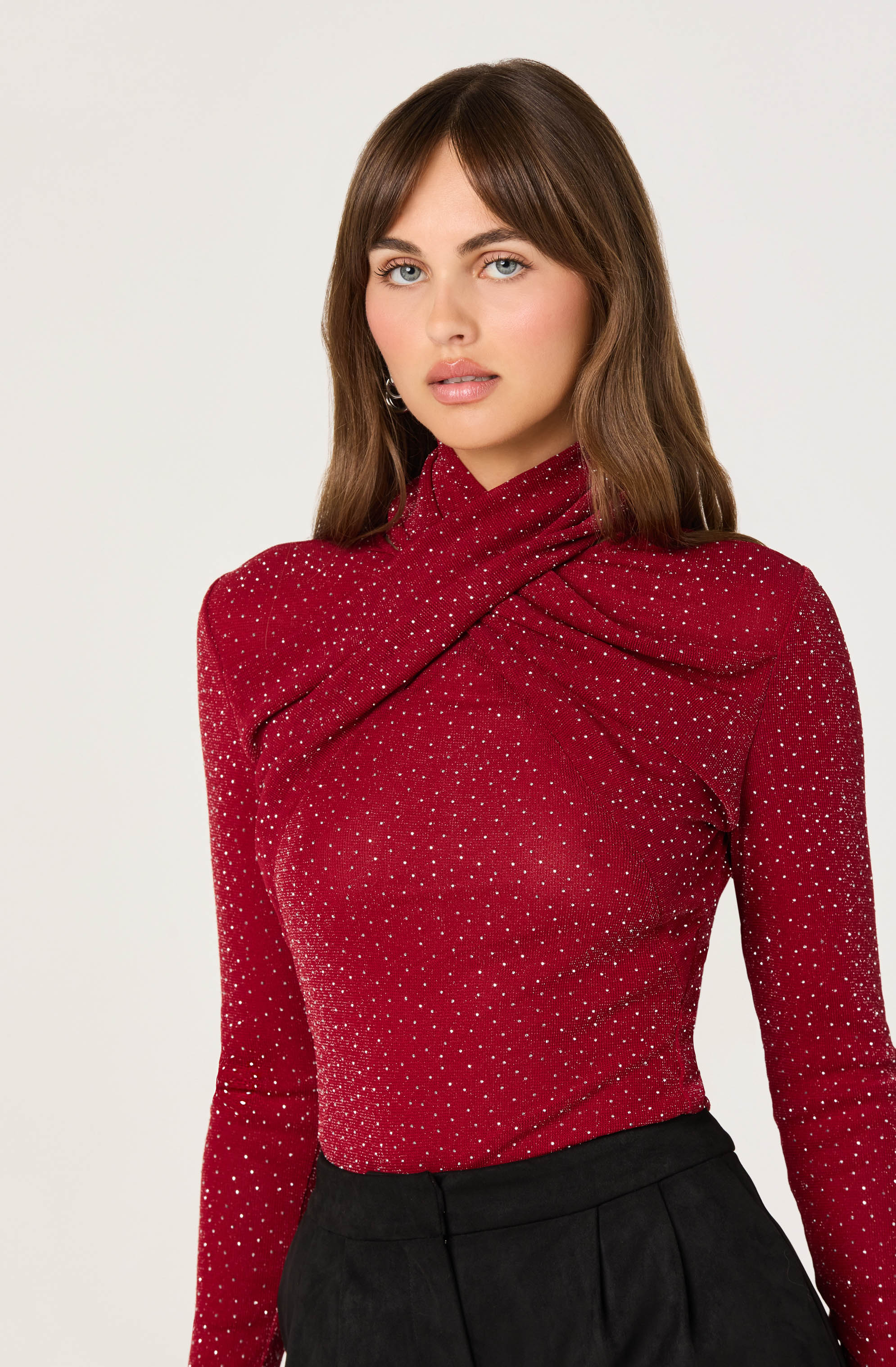 A woman is wearing a fitted, long-sleeved red top with a high, asymmetrical neckline that wraps around her neck. The top is adorned with small, evenly spaced, white polka dots. She has paired the top with high-waisted black pants.