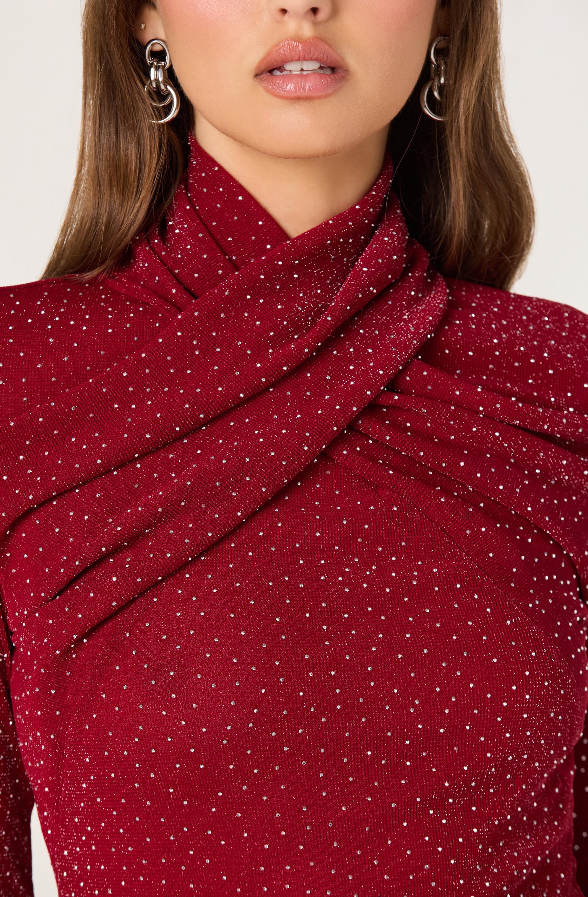 A woman is wearing a red, glittery fabric top with a high neckline that drapes elegantly across her shoulders. The top is decorated with numerous small, sparkling silver dots that create a shimmering effect throughout the material.