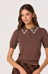 A woman is wearing a brown, short-sleeved knit top with a collar decorated with white floral embroidery. The top has a ribbed band at the hem and a button placket at the neckline. She is also wearing a pair of high-waisted plaid pants in shades of brown and orange.